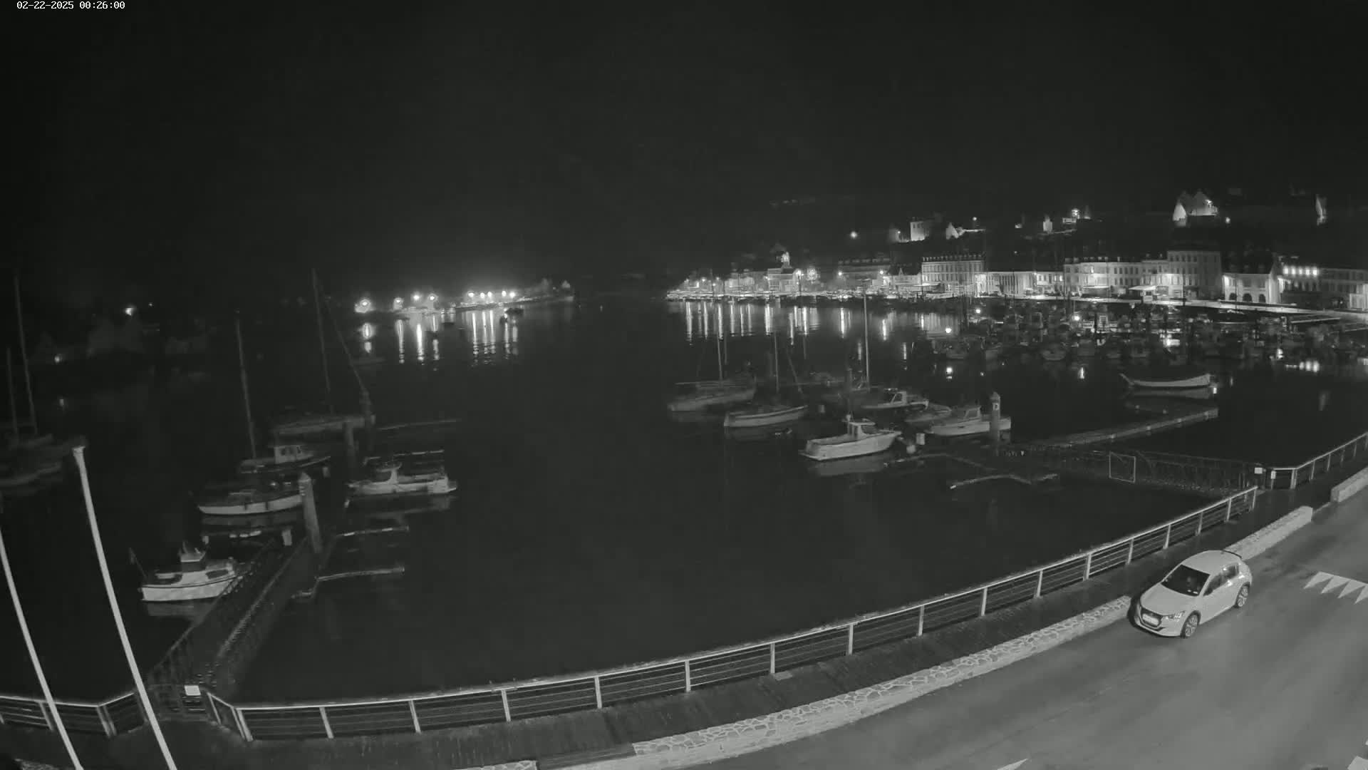 A nighttime view of a harbor filled with boats, alongside a waterfront town with lights reflecting on the calm water, under a clear, dark sky.