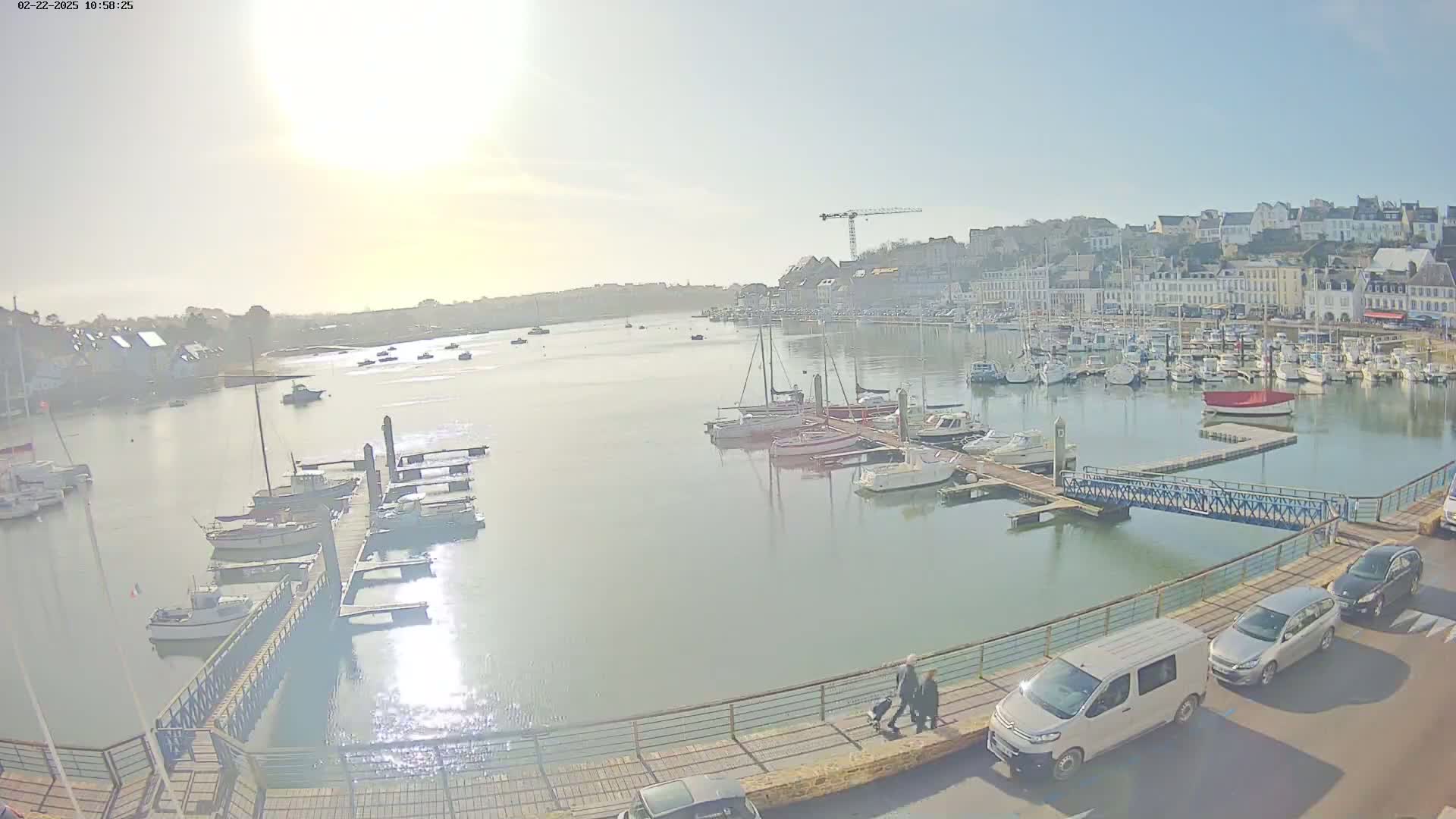 A sunny harbor scene shows numerous boats docked at piers, with a town in the background and a few pedestrians walking along a waterfront walkway.
