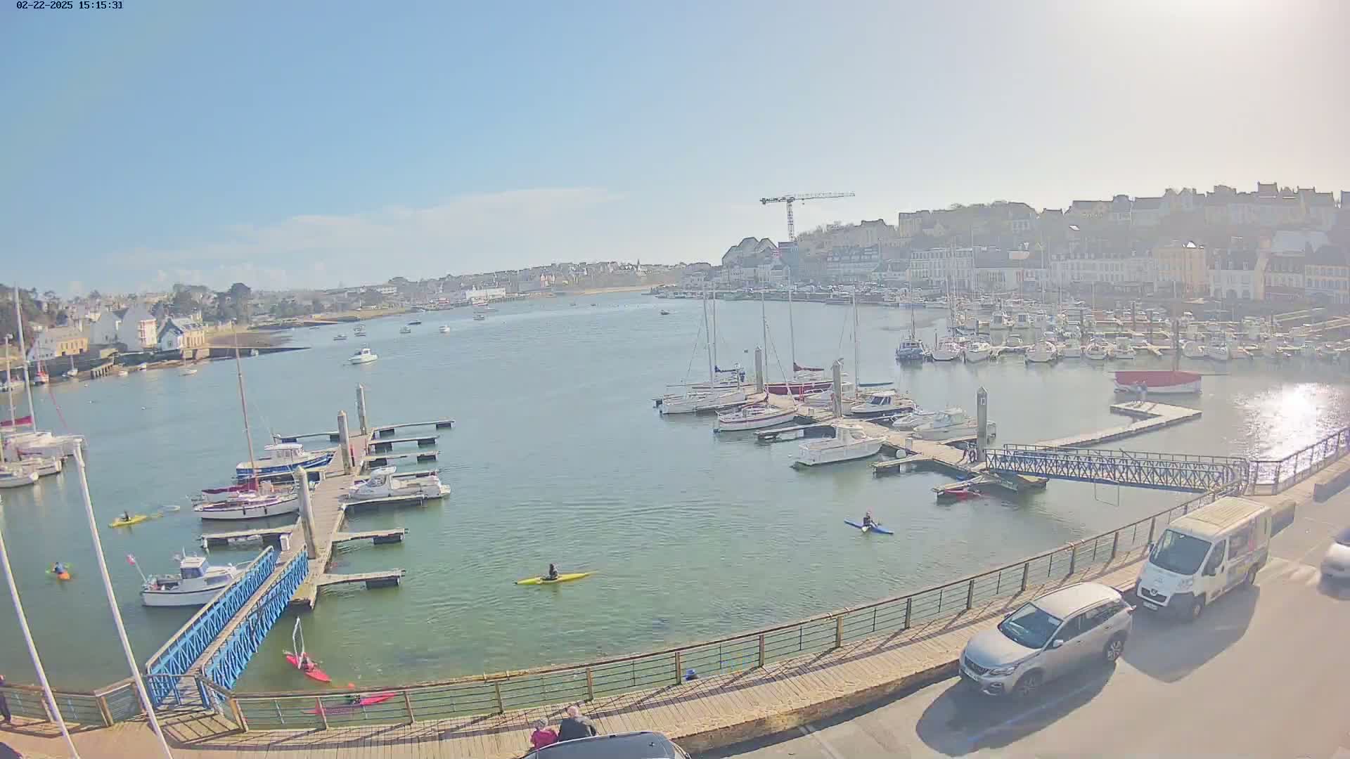 A sunny day at a harbor shows numerous boats docked at piers, with kayakers on the water and cars parked nearby, all overlooking a town.