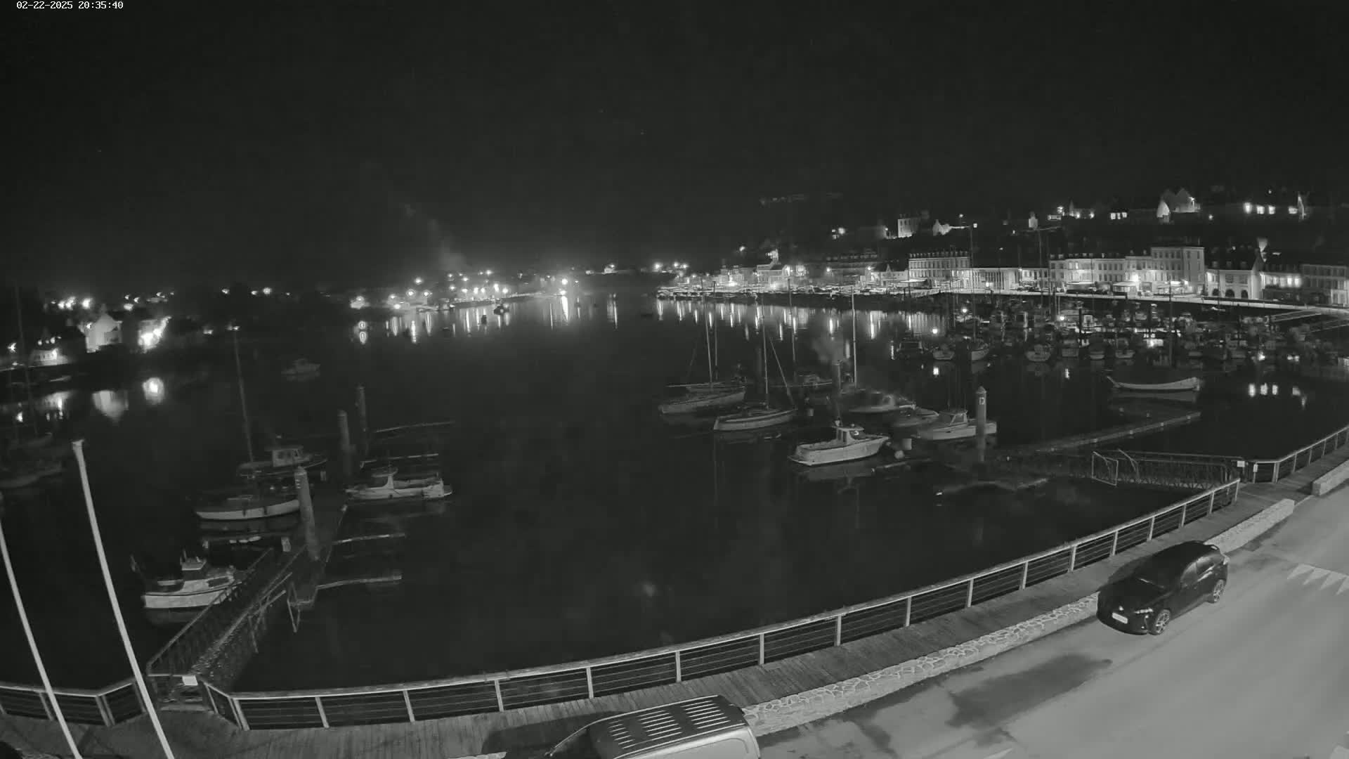 A nighttime, high-angle view shows a harbor filled with boats and a town with lights reflecting on the water.