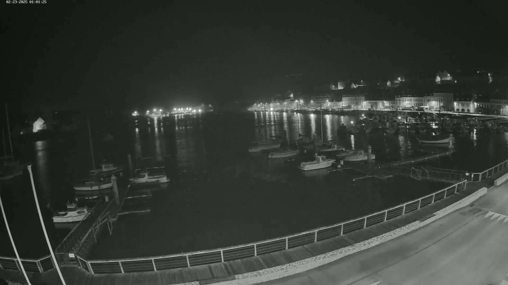 A nighttime view of a harbor filled with boats, with a town's lights reflecting on the calm water.