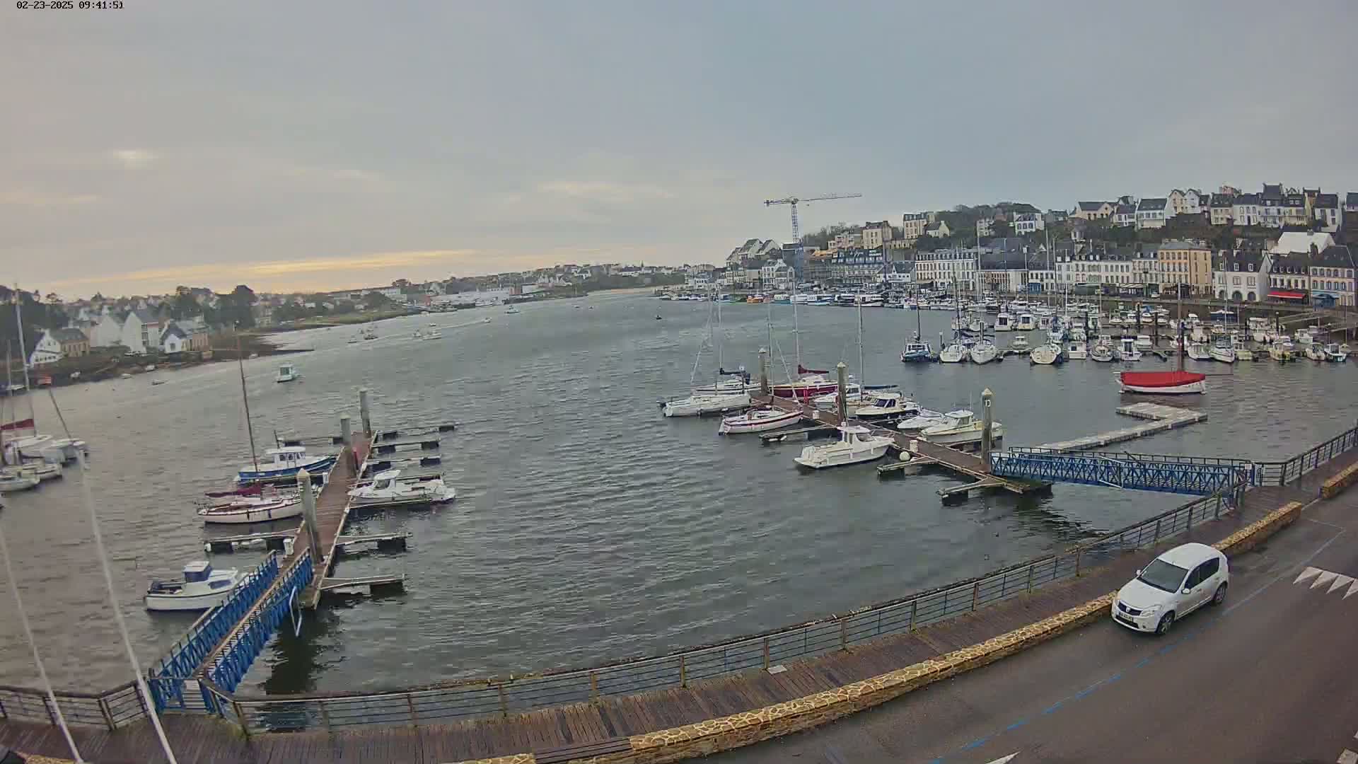 A harbor filled with numerous boats and yachts is seen under an overcast sky, with a town and a crane visible in the background.