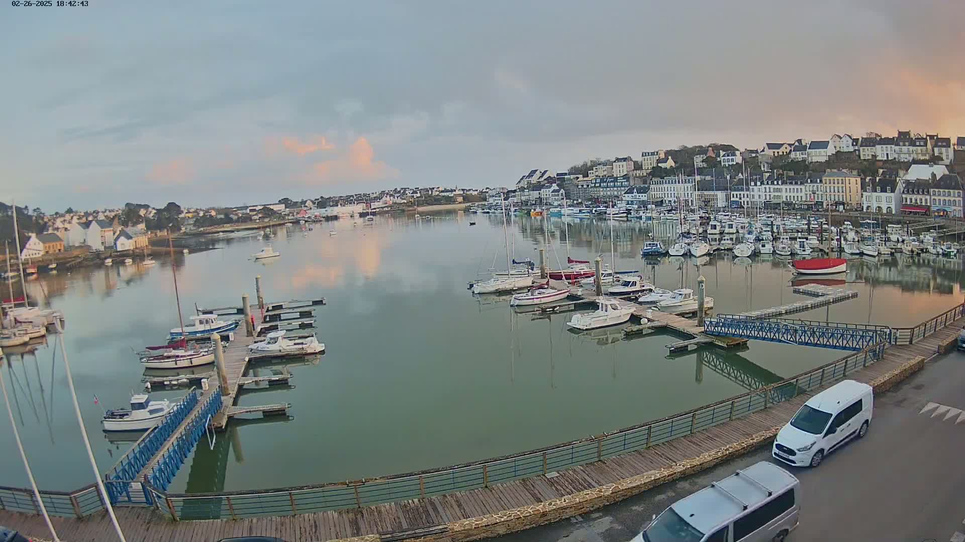 A calm harbor filled with numerous boats and yachts is nestled beside a town under a partly cloudy, twilight sky.
