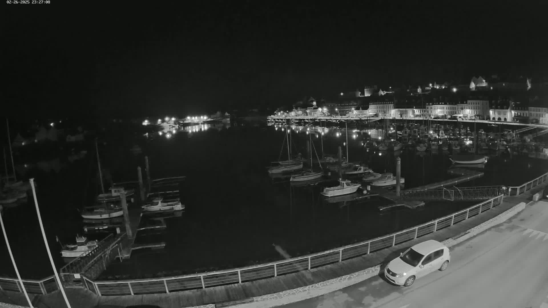A nighttime harbor scene shows numerous boats docked in calm water, with a town's lights reflected on the water and a single car parked on a nearby road.