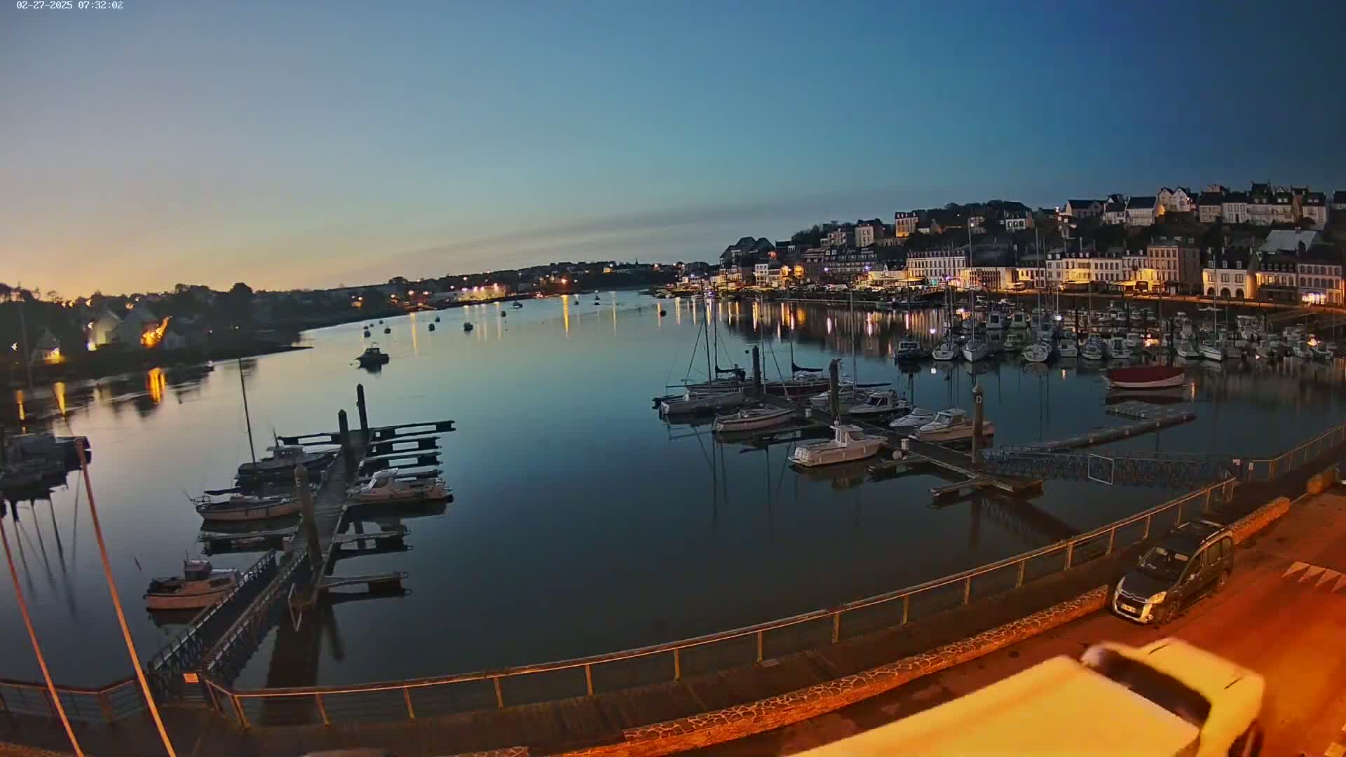 A calm body of water at dawn, filled with numerous boats docked at a marina, overlooks a town with buildings illuminated along the shore.