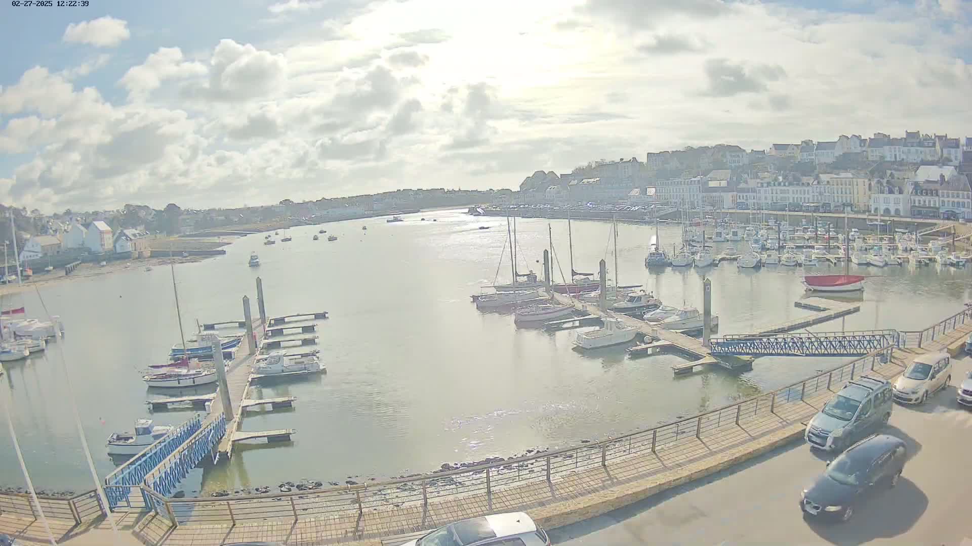 A harbor filled with numerous boats and yachts under a partly cloudy sky, with cars parked along a nearby road.