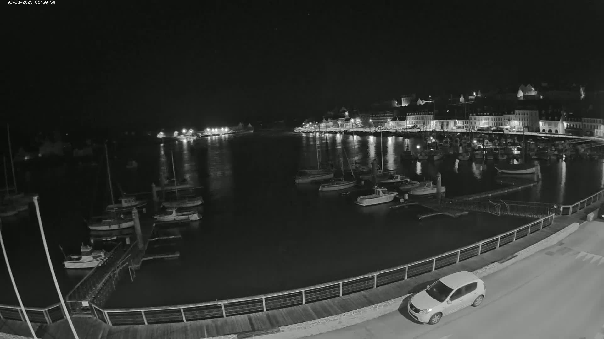 A nighttime, low-light view of a harbor filled with docked boats, a small car parked on a nearby road, and a town's lights visible in the background.