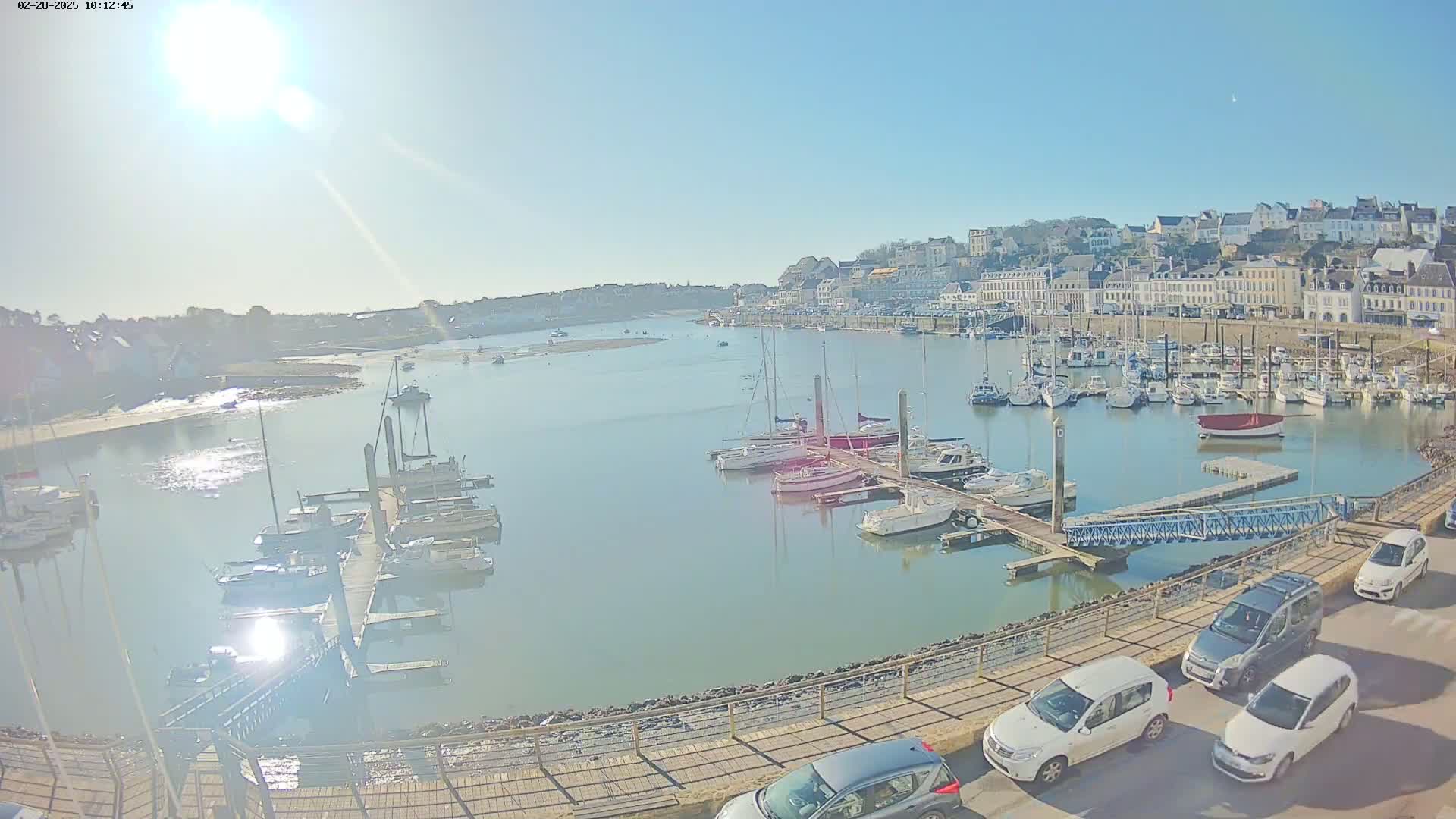 A sunny day overlooking a harbor filled with sailboats and motorboats, a town in the background, and several cars parked along a walkway.