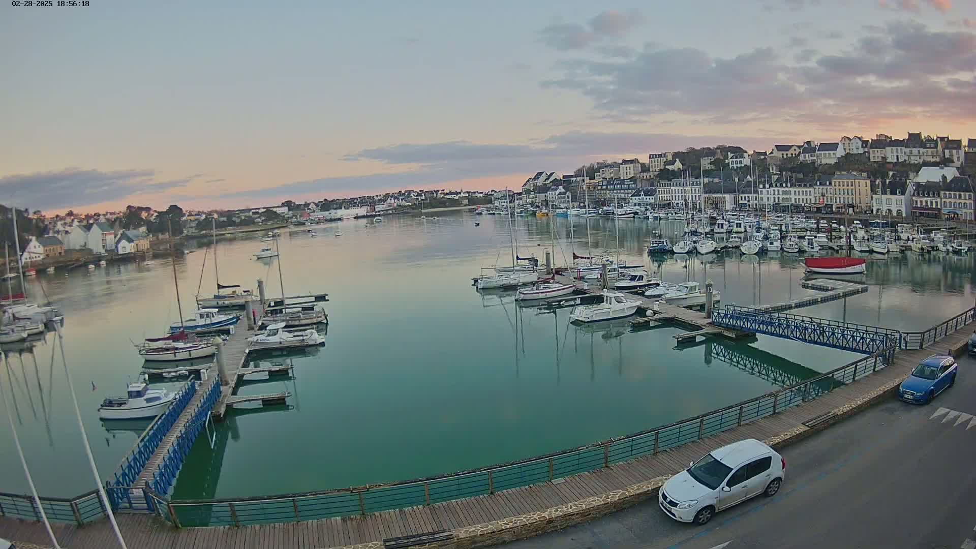 A calm harbor filled with numerous sailboats and motorboats at sunset, overlooked by a town of white buildings.