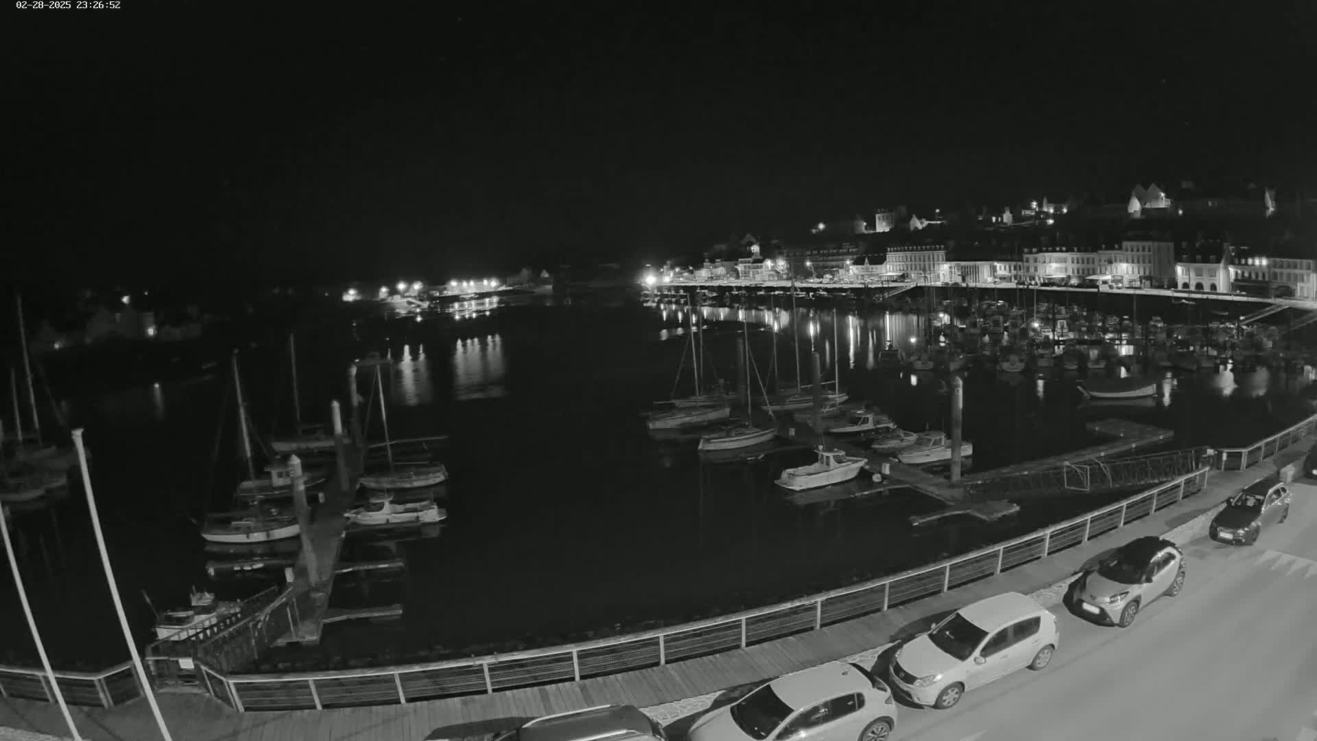 A nighttime, black-and-white view of a harbor filled with docked sailboats and small motorboats, with cars parked along a waterfront walkway and buildings illuminated in the distance.
