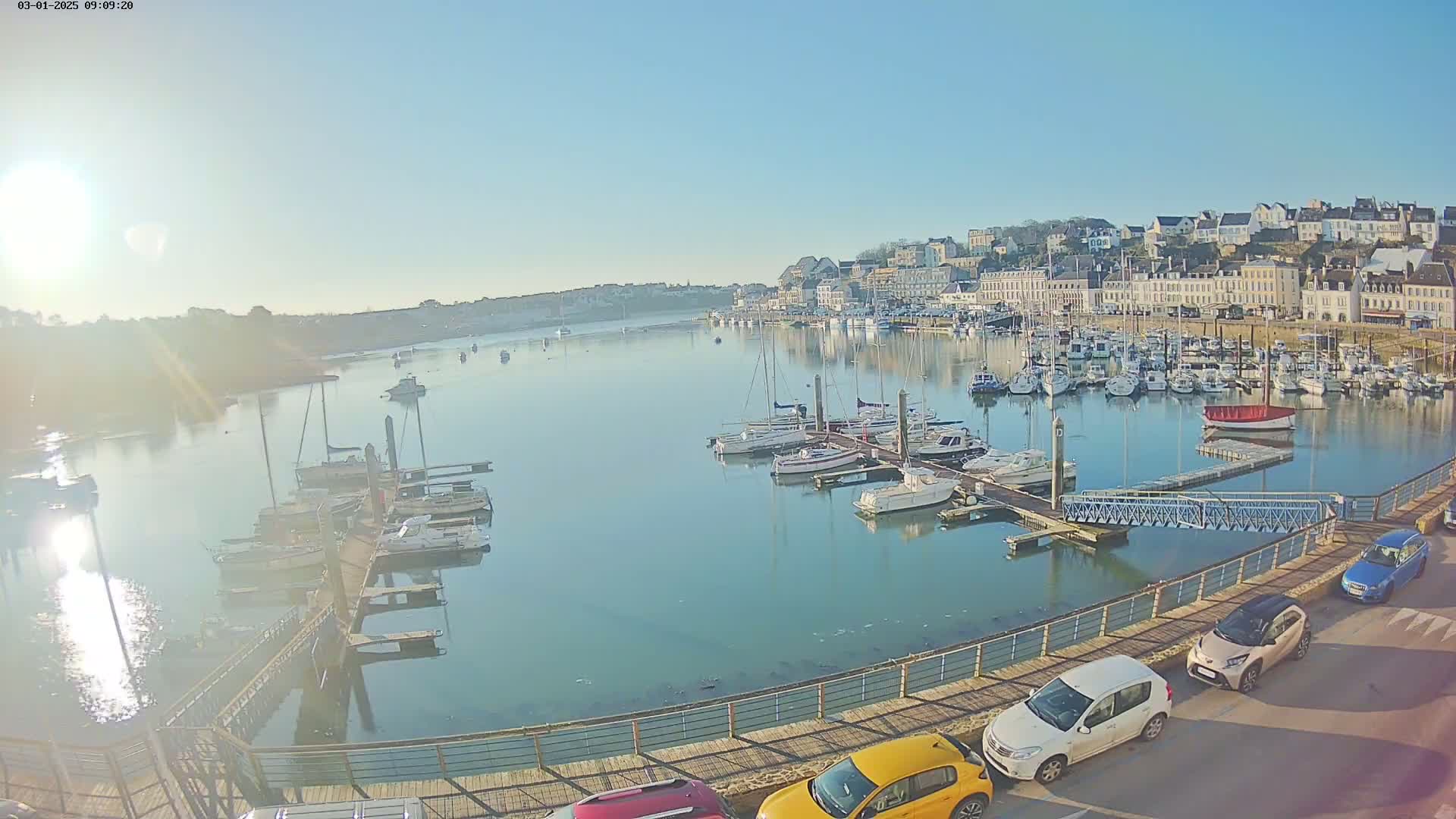A harbor filled with numerous sailboats and motorboats under a clear, sunny sky, with a town visible in the background and cars parked along a walkway.