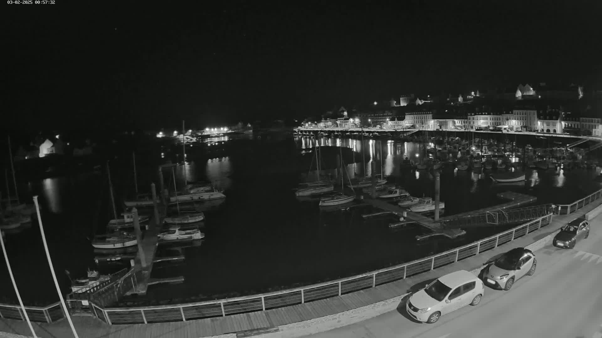 A nighttime, black-and-white view of a harbor filled with boats, a town in the background, and two cars parked near a railing.