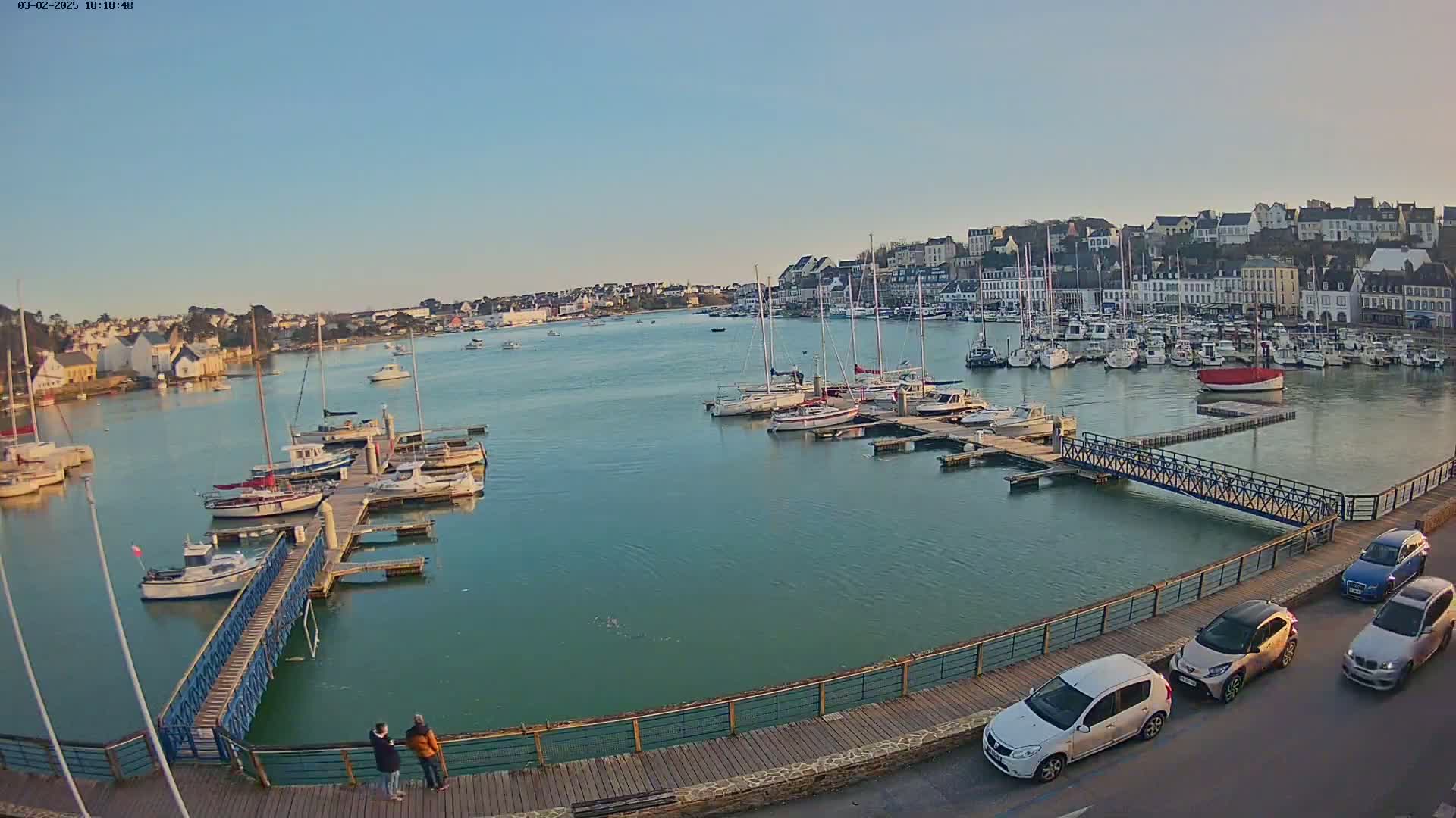 A calm harbor filled with sailboats and motorboats, under a clear sky, is viewed from a boardwalk with several cars parked along the side.