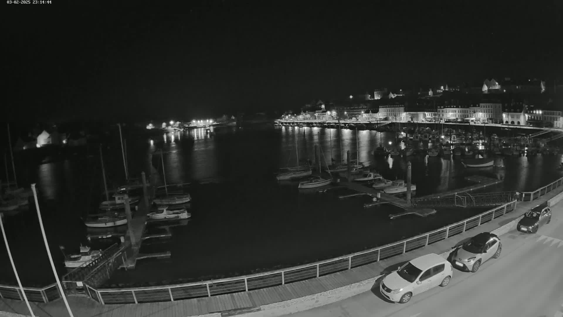 A nighttime view of a harbor filled with boats, alongside a waterfront town illuminated with lights under a clear, dark sky.