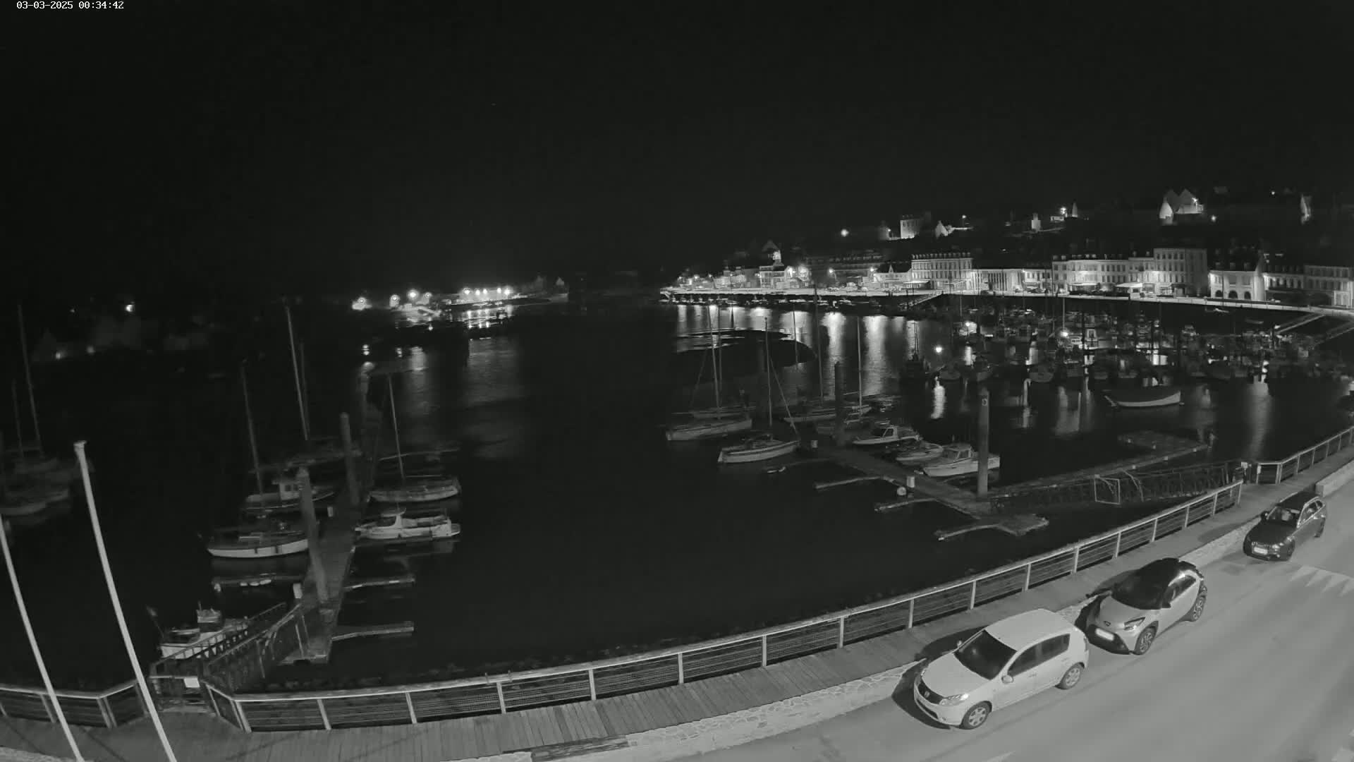 A nighttime view of a harbor filled with docked boats, a town's waterfront buildings, and two cars parked alongside a walkway.