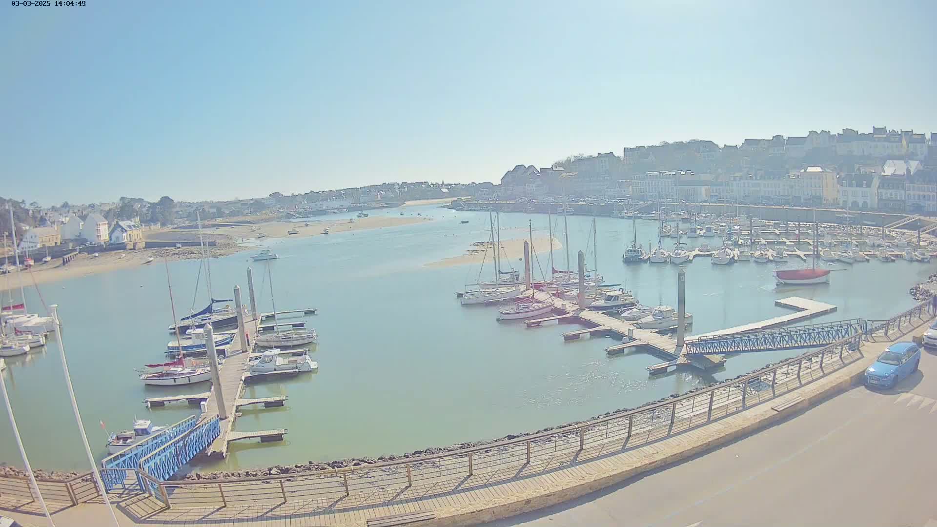 A marina filled with sailboats and motorboats is situated in front of a town under a clear, sunny sky.