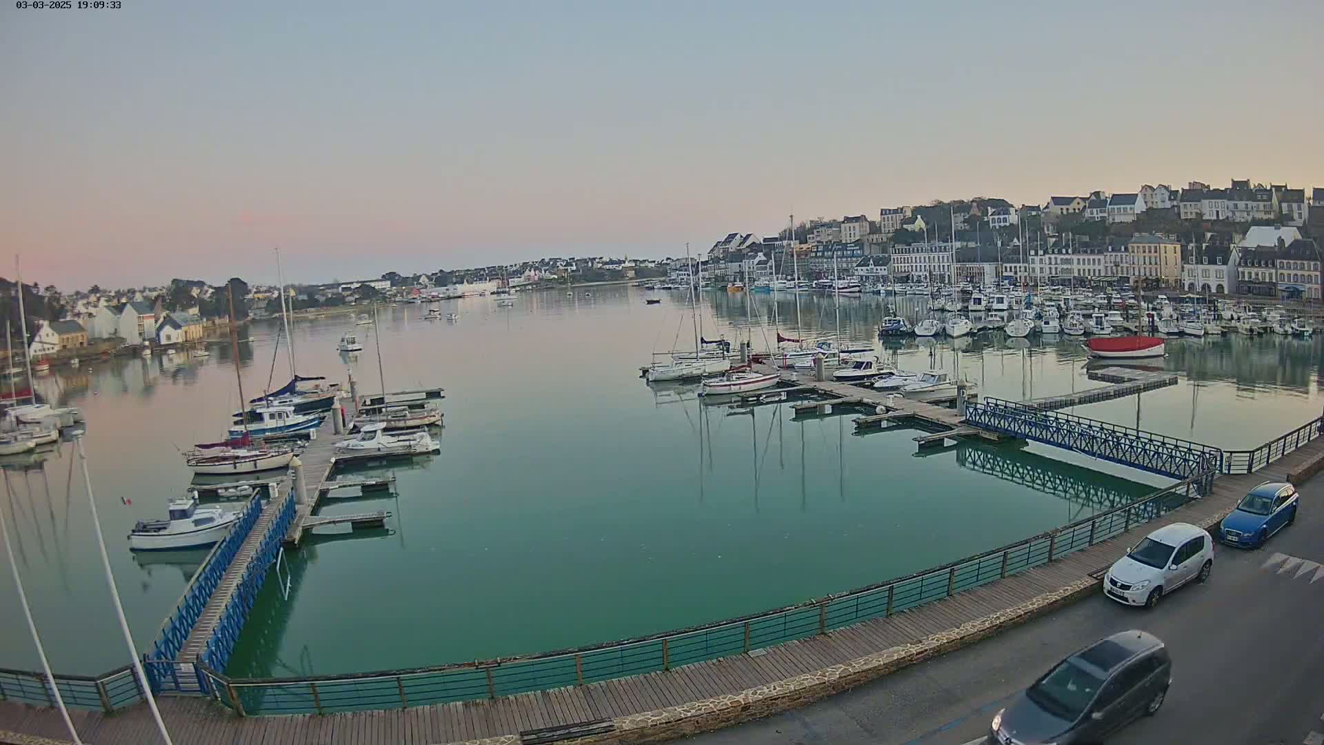 A calm harbor filled with numerous sailboats and motorboats at dusk, next to a waterfront town.