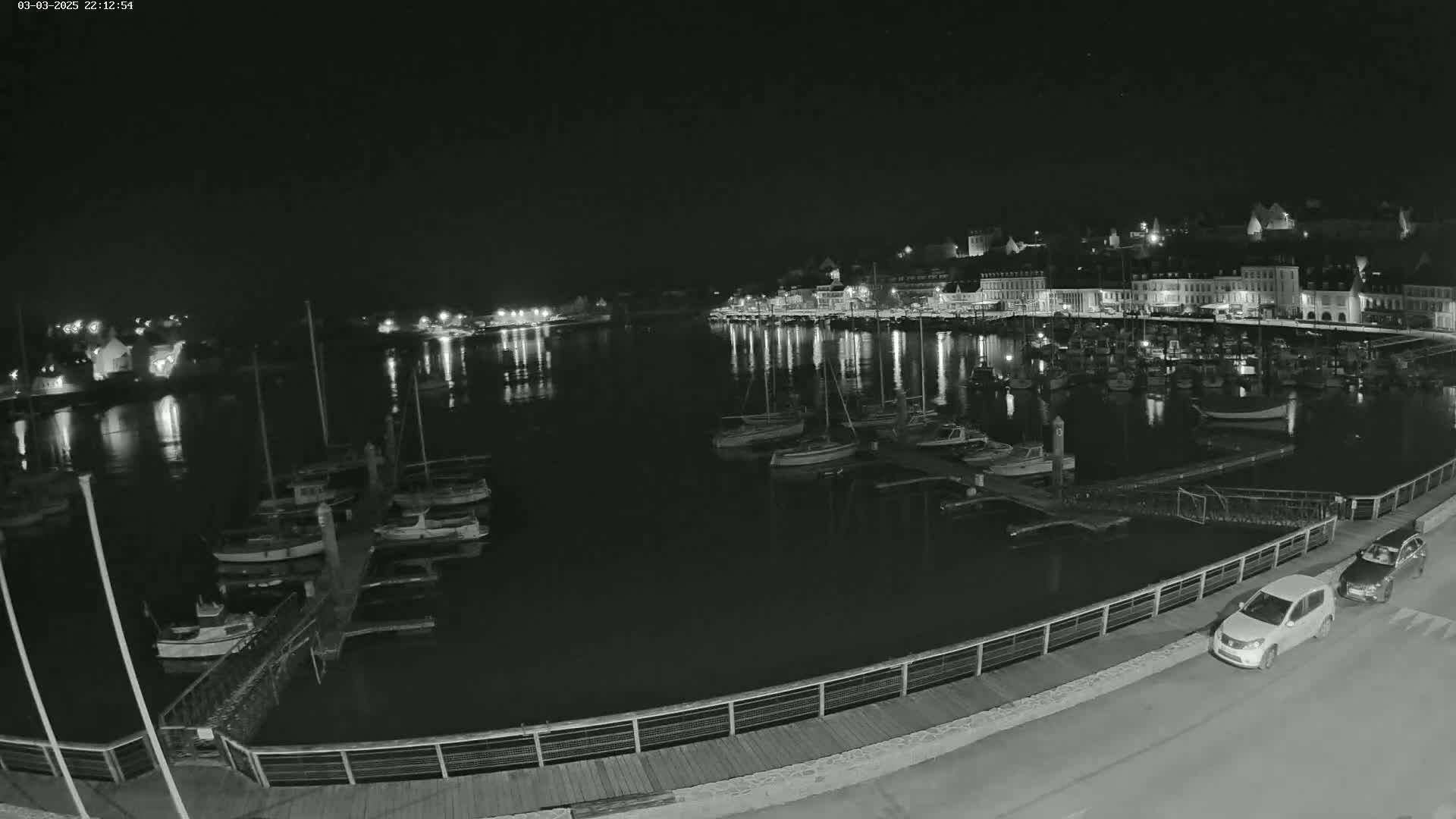 A nighttime view of a harbor filled with docked boats, a waterfront town with lights reflecting on the water, and two cars parked along a nearby road.