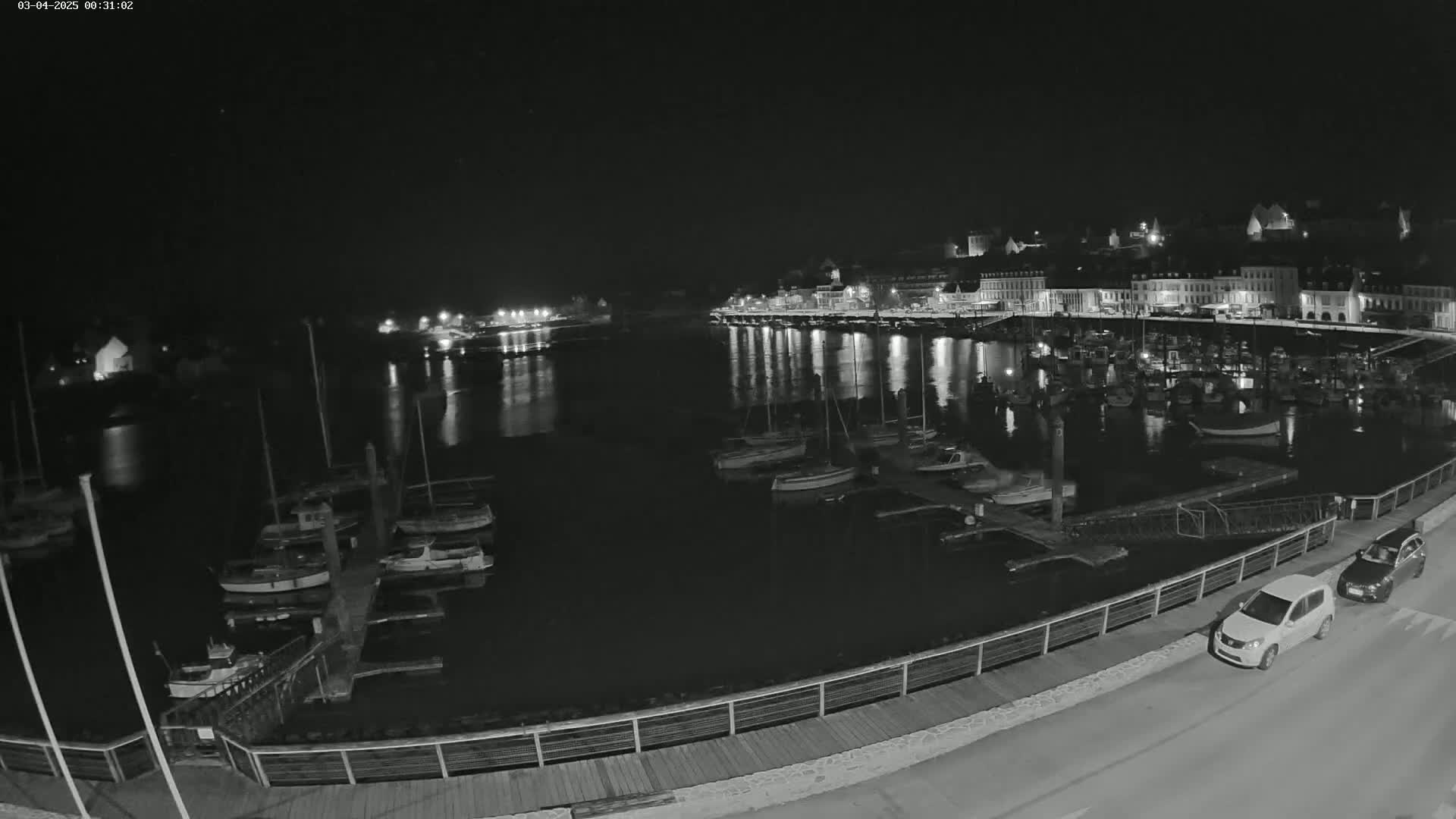 A nighttime view of a harbor filled with docked boats, with a town's lights reflecting on the calm water.