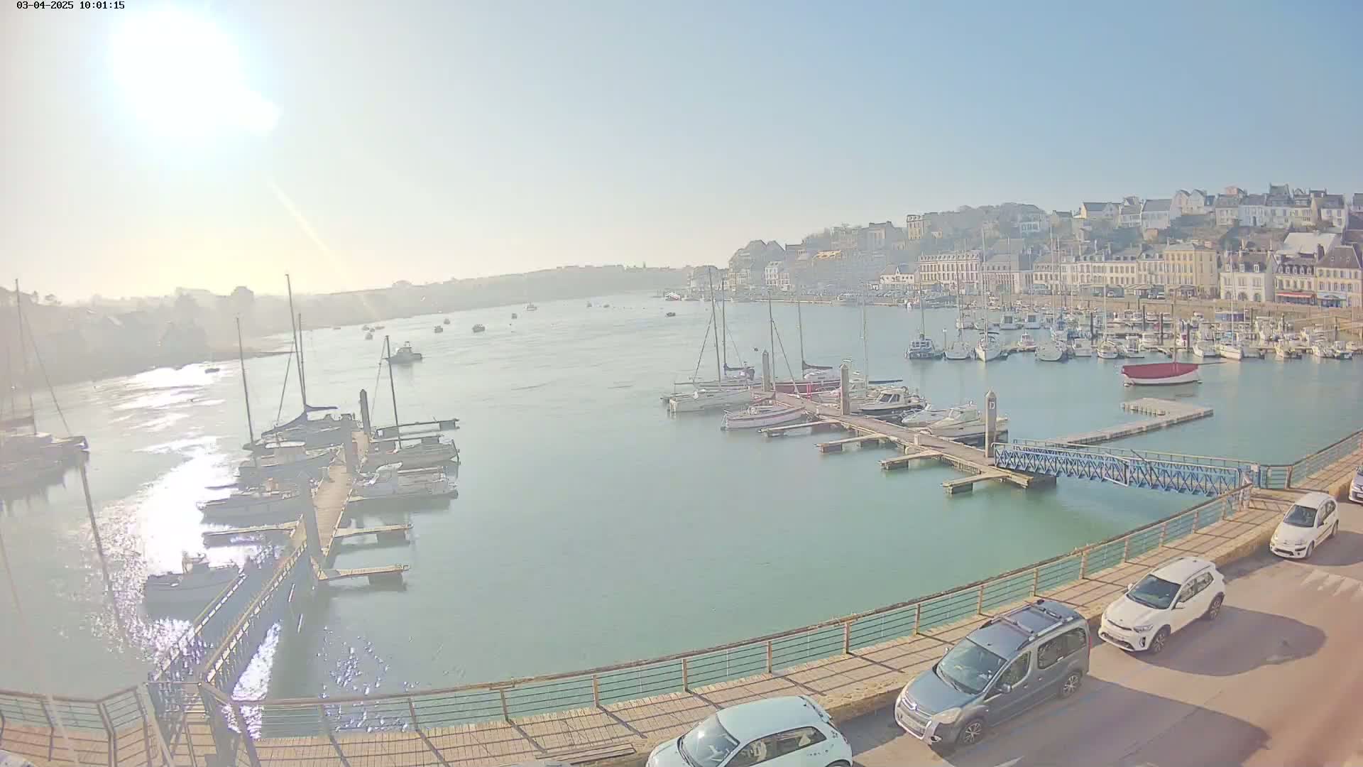 A harbor filled with sailboats and motorboats on a sunny day, with a town visible in the background and several cars parked along a walkway.