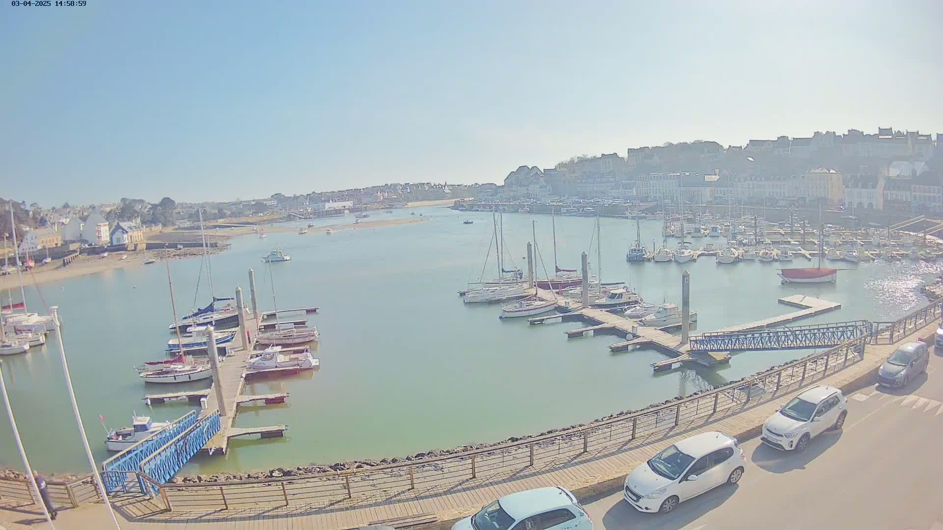 A harbor filled with sailboats and motorboats on a sunny day, with a town visible in the background and several cars parked along a paved walkway.