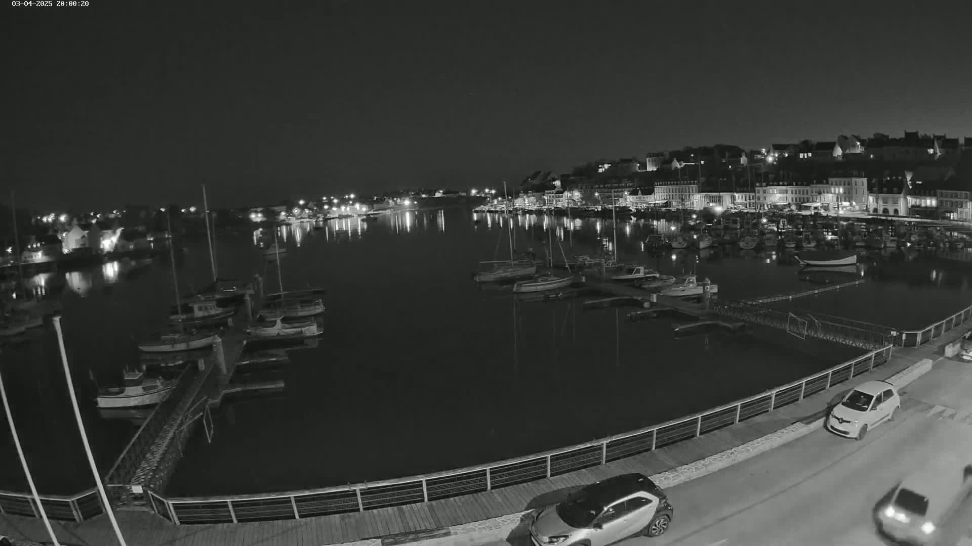 A nighttime harbor scene shows numerous boats docked along a waterfront, with a town's illuminated buildings visible in the background under a clear, dark sky.