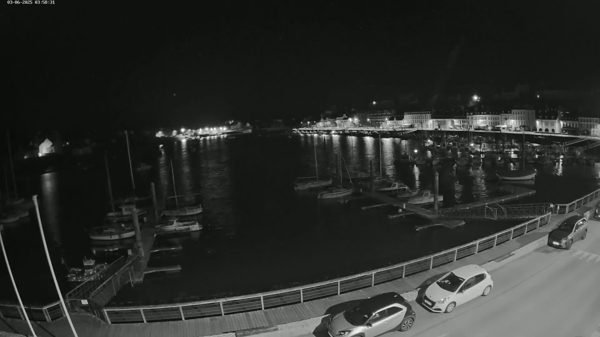 A nighttime, low-light view of a harbor filled with docked boats, next to a waterfront with cars parked along the road.