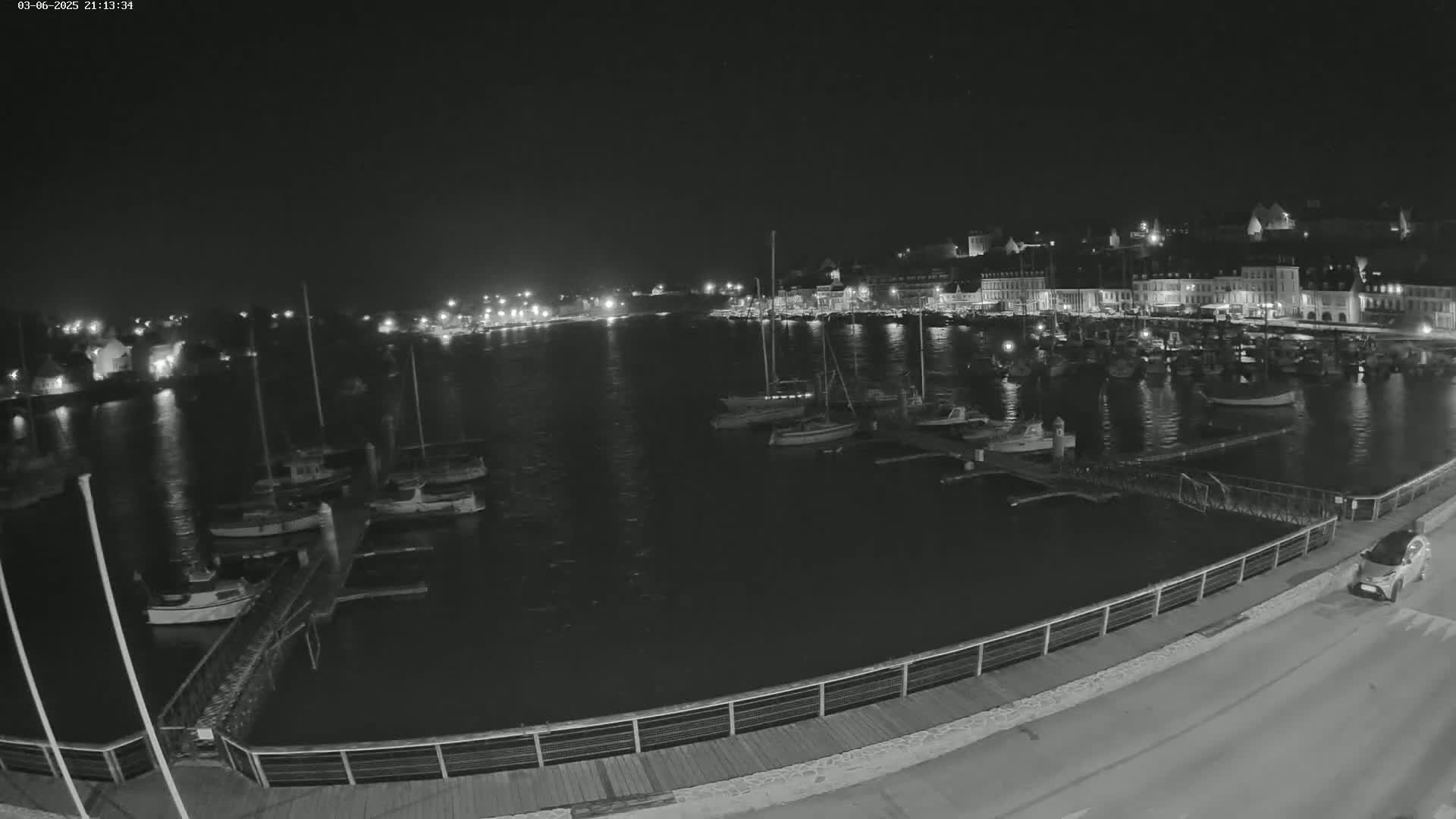 A nighttime, harbor scene shows numerous boats docked in calm water, with a town's lights illuminating the shore in the background.