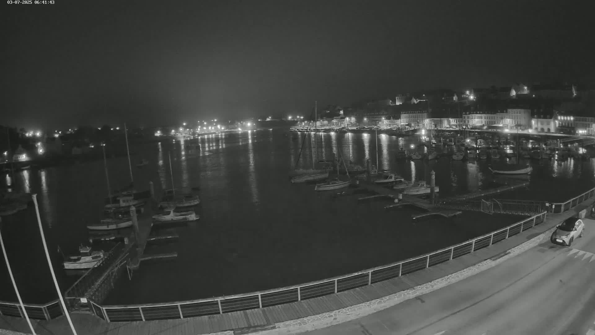 A nighttime, black and white view of a harbor filled with boats, a town's lights reflecting in the calm water, and a car driving along a nearby road.
