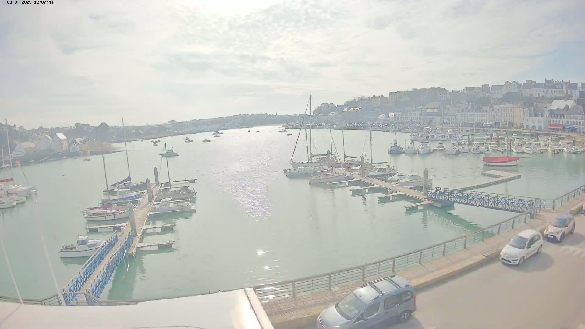 A harbor filled with numerous boats and yachts under a partly cloudy sky, with a town visible in the background.