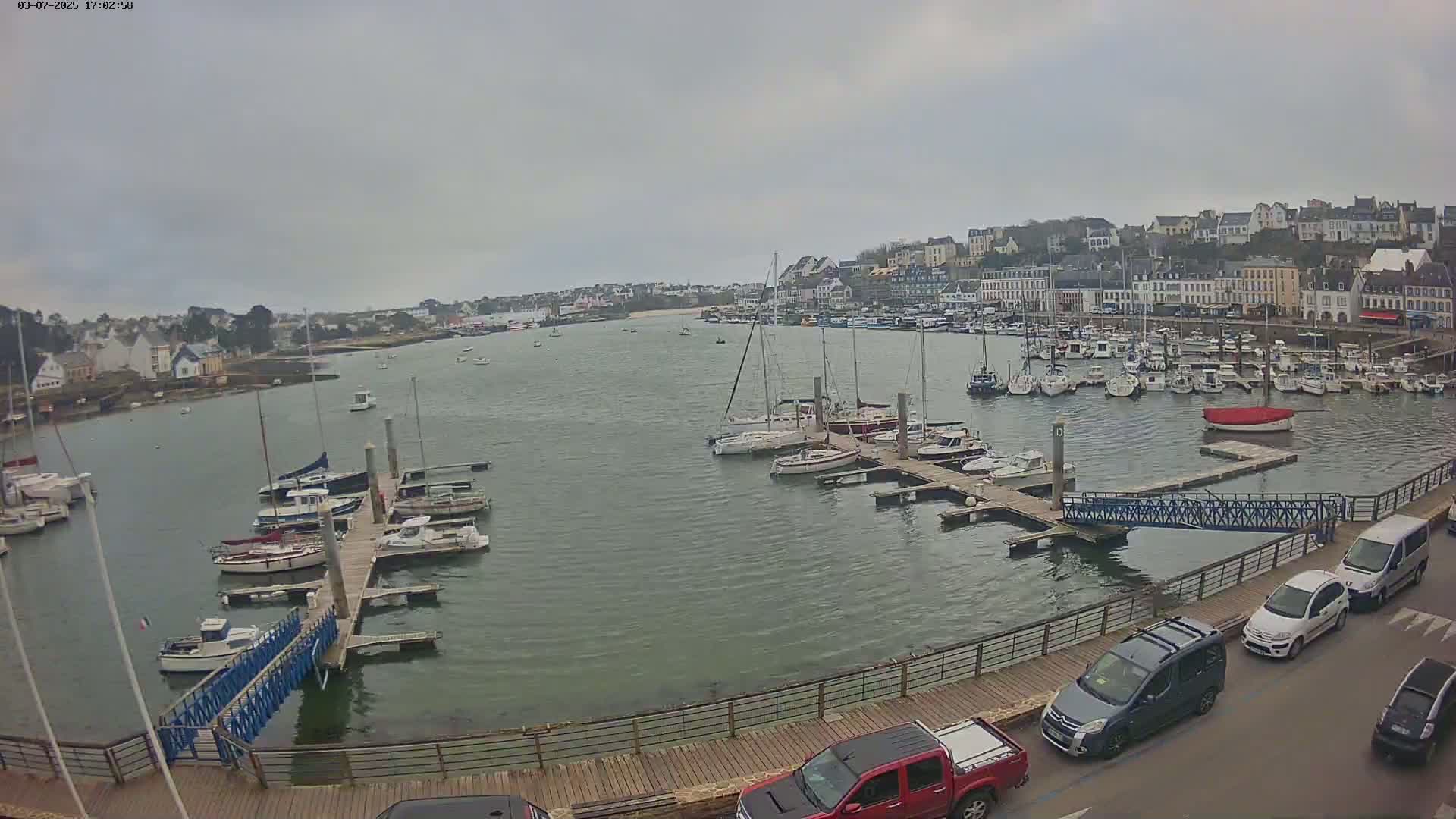 A harbor filled with numerous boats and yachts under an overcast sky, with a town visible in the background and cars parked along a nearby roadway.