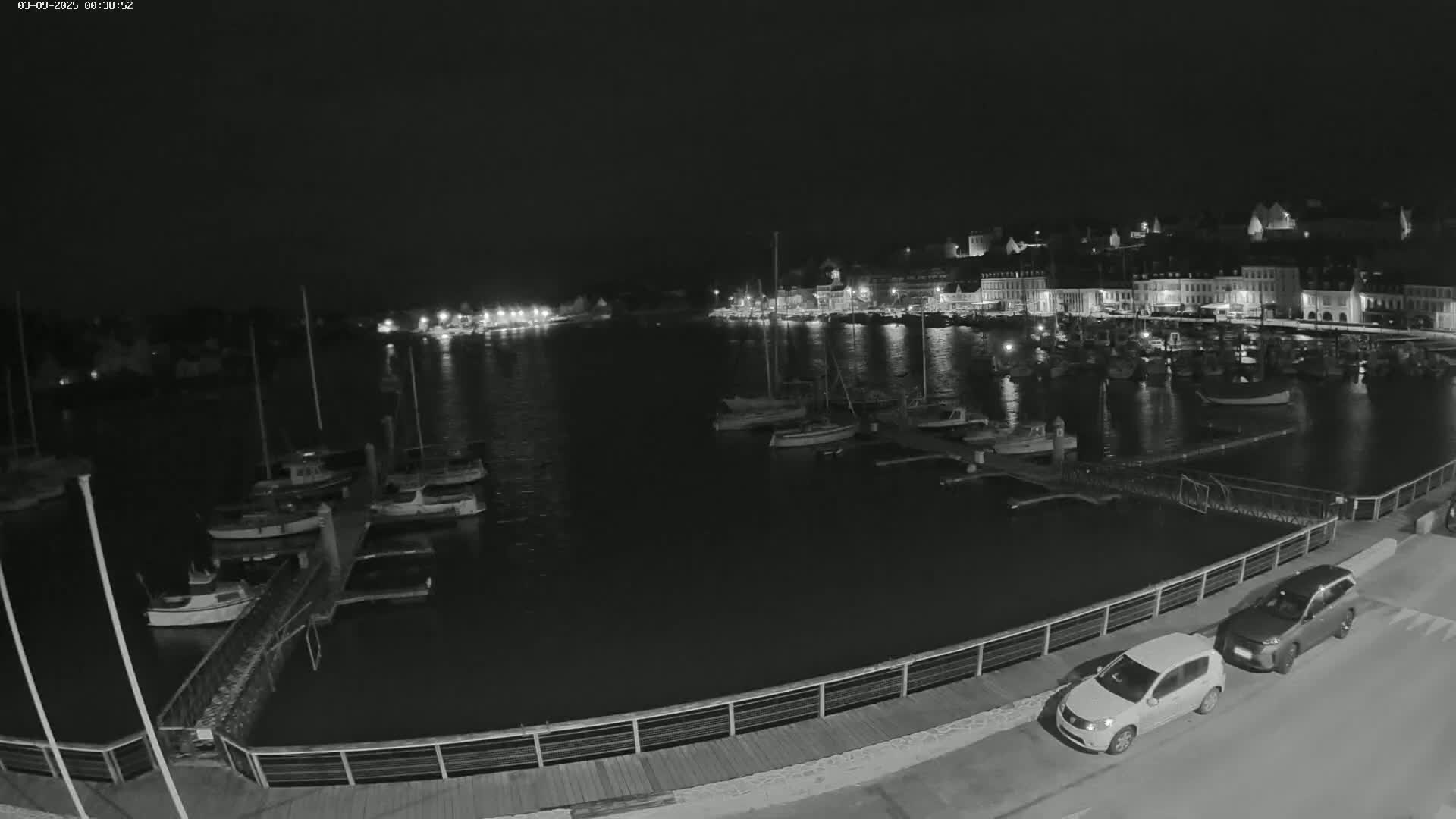 A nighttime view of a harbor filled with boats, with a town illuminated in the background, and two cars parked on a nearby road.