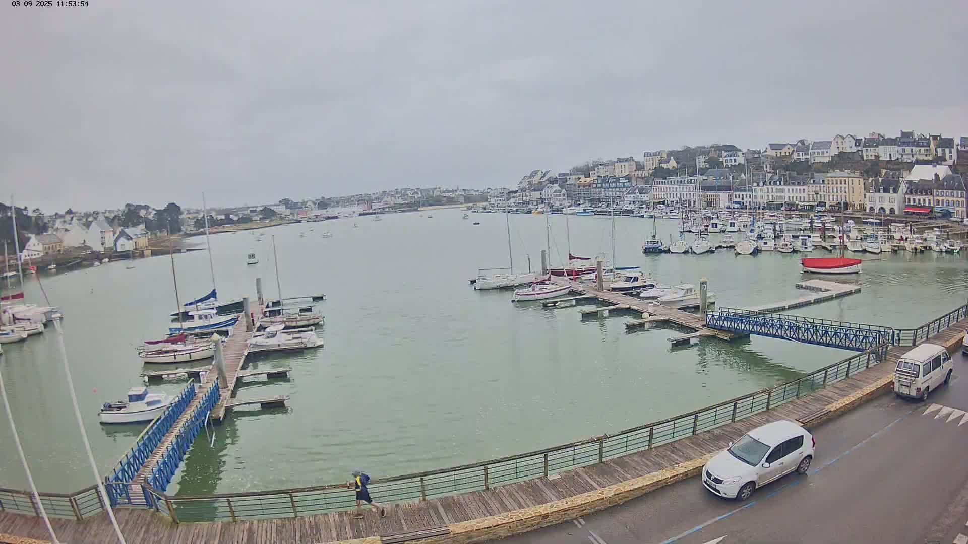 A harbor filled with sailboats and motorboats on a cloudy day, with a person jogging along a boardwalk next to a road with cars.