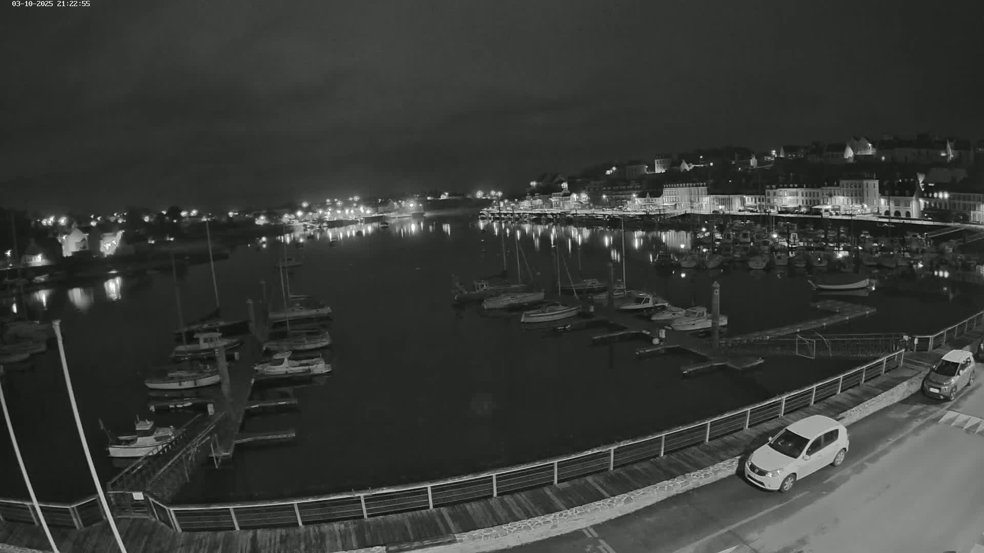 A nighttime, black-and-white view of a harbor filled with boats, alongside a waterfront town with lights reflecting on the water.