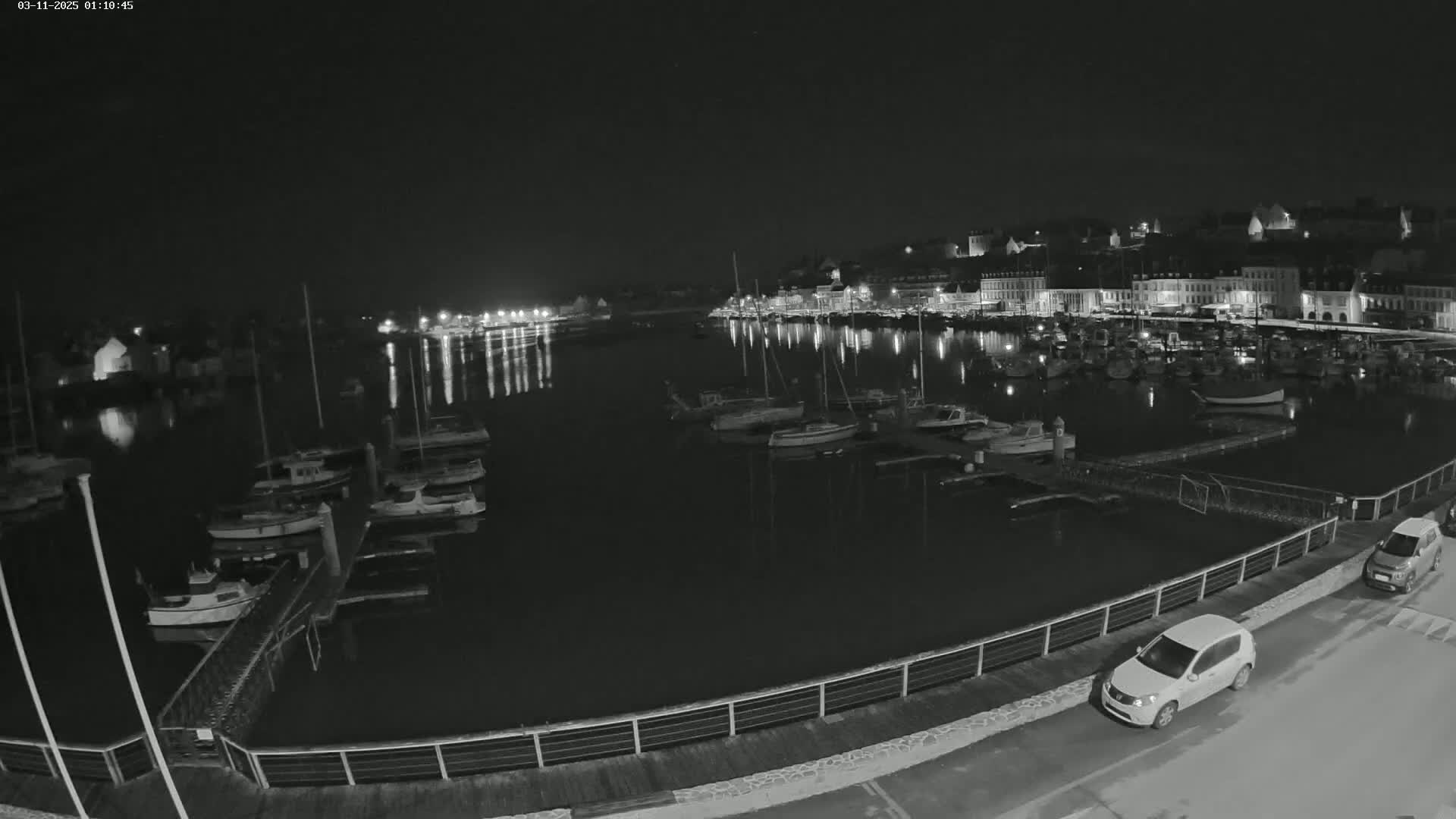 A nighttime view of a harbor filled with boats, docked along a waterfront with buildings and streetlights visible in the background under a clear, dark sky.