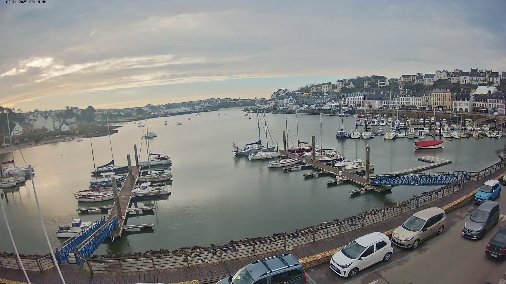 A harbor filled with sailboats and motorboats under an overcast sky, with a town visible in the background and cars parked along a waterfront walkway.