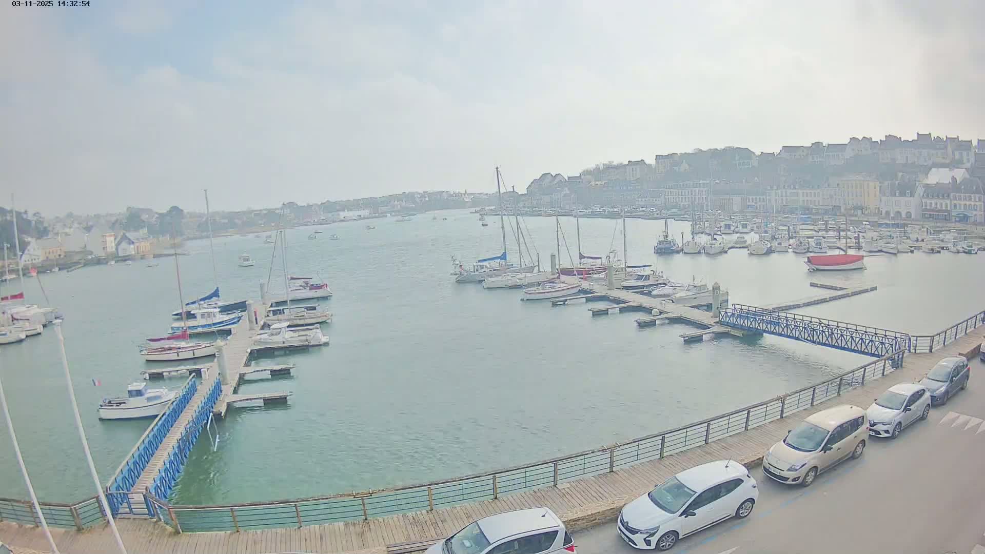 A harbor filled with sailboats and motorboats on a cloudy day, with a town and cars parked along a boardwalk in the foreground.