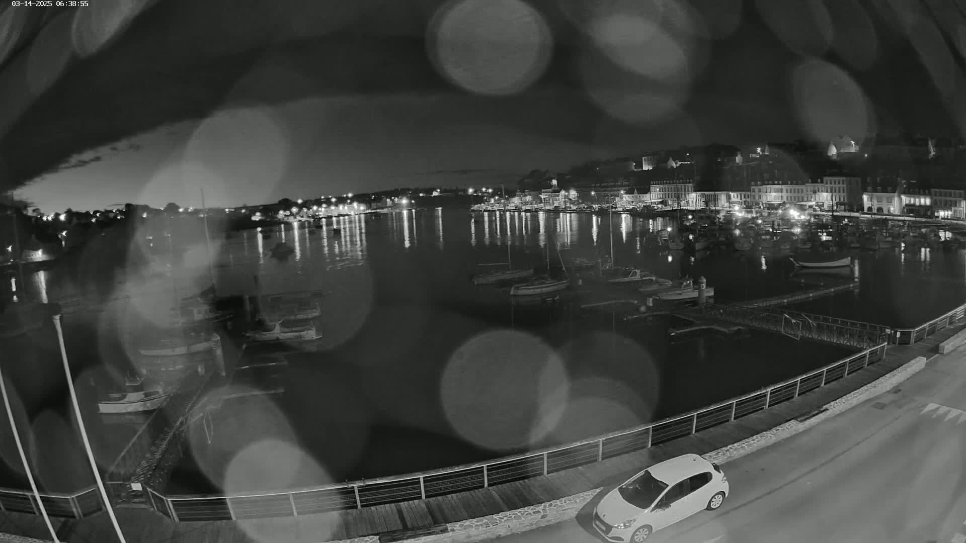 A nighttime, rain-blurred view of a harbor filled with boats, overlooked by a town with lights reflecting on the water, and a car parked on a nearby road.