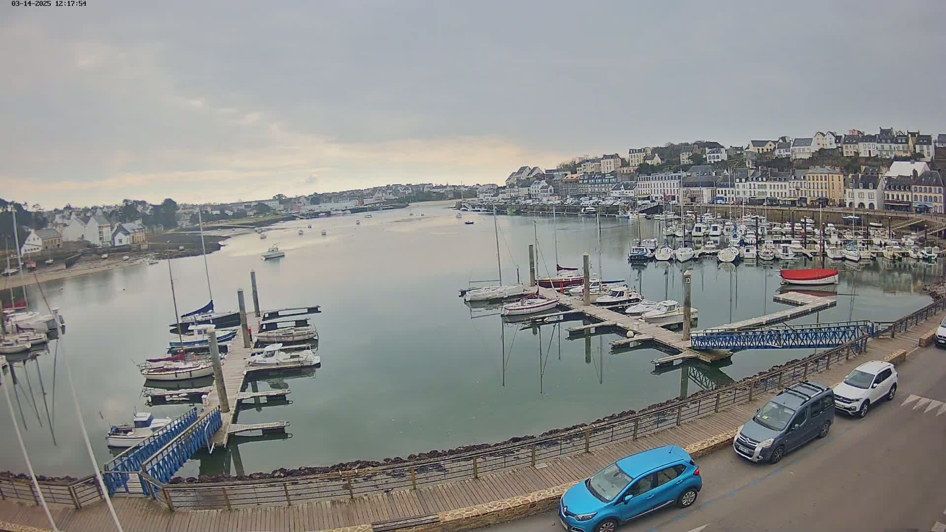 A harbor filled with sailboats and motorboats under an overcast sky, with a town in the background and cars parked along a walkway.