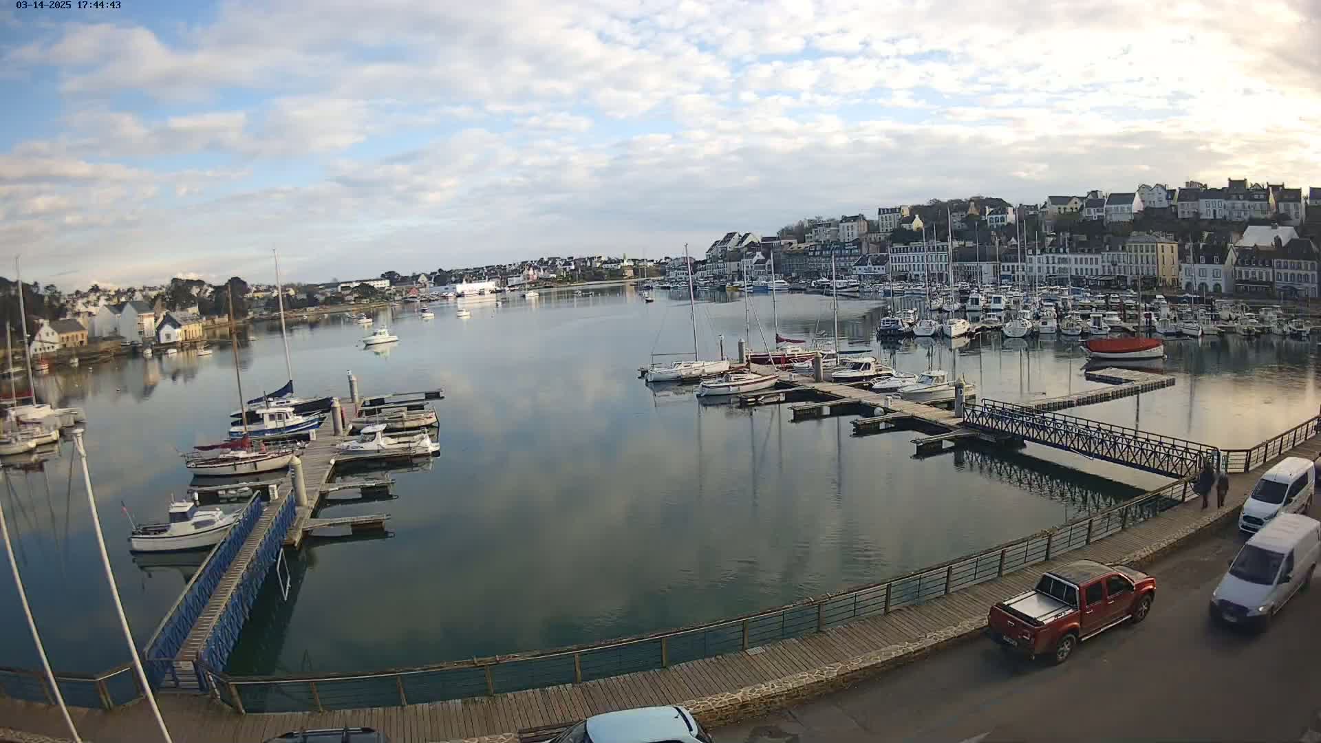 A harbor filled with numerous sailboats and motorboats is seen on a partly cloudy day, with a town's buildings in the background.