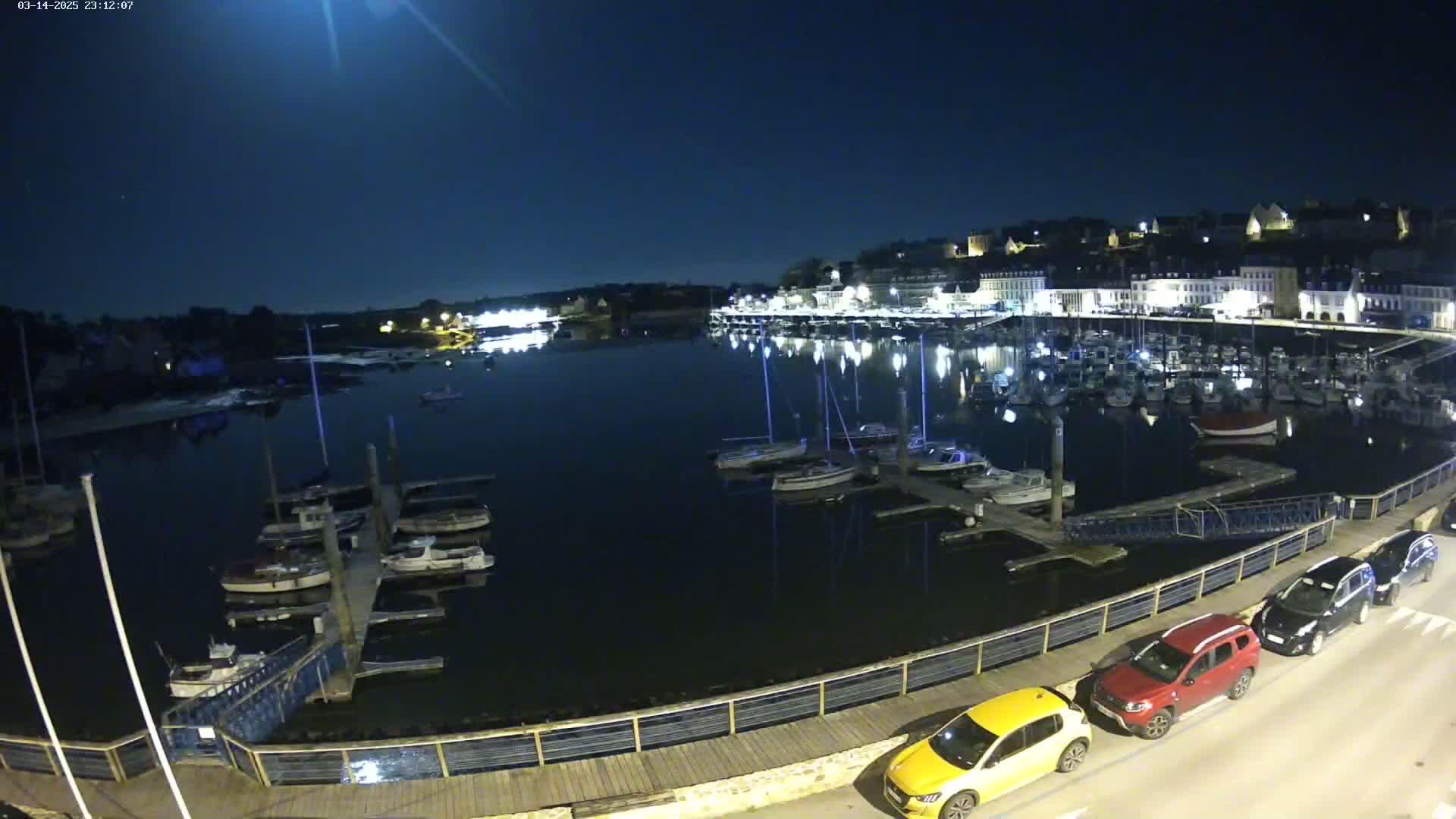 A nighttime view of a harbor filled with boats, a town with lights reflecting in the water, and a few cars parked along a waterfront walkway under a clear, moonlit sky.