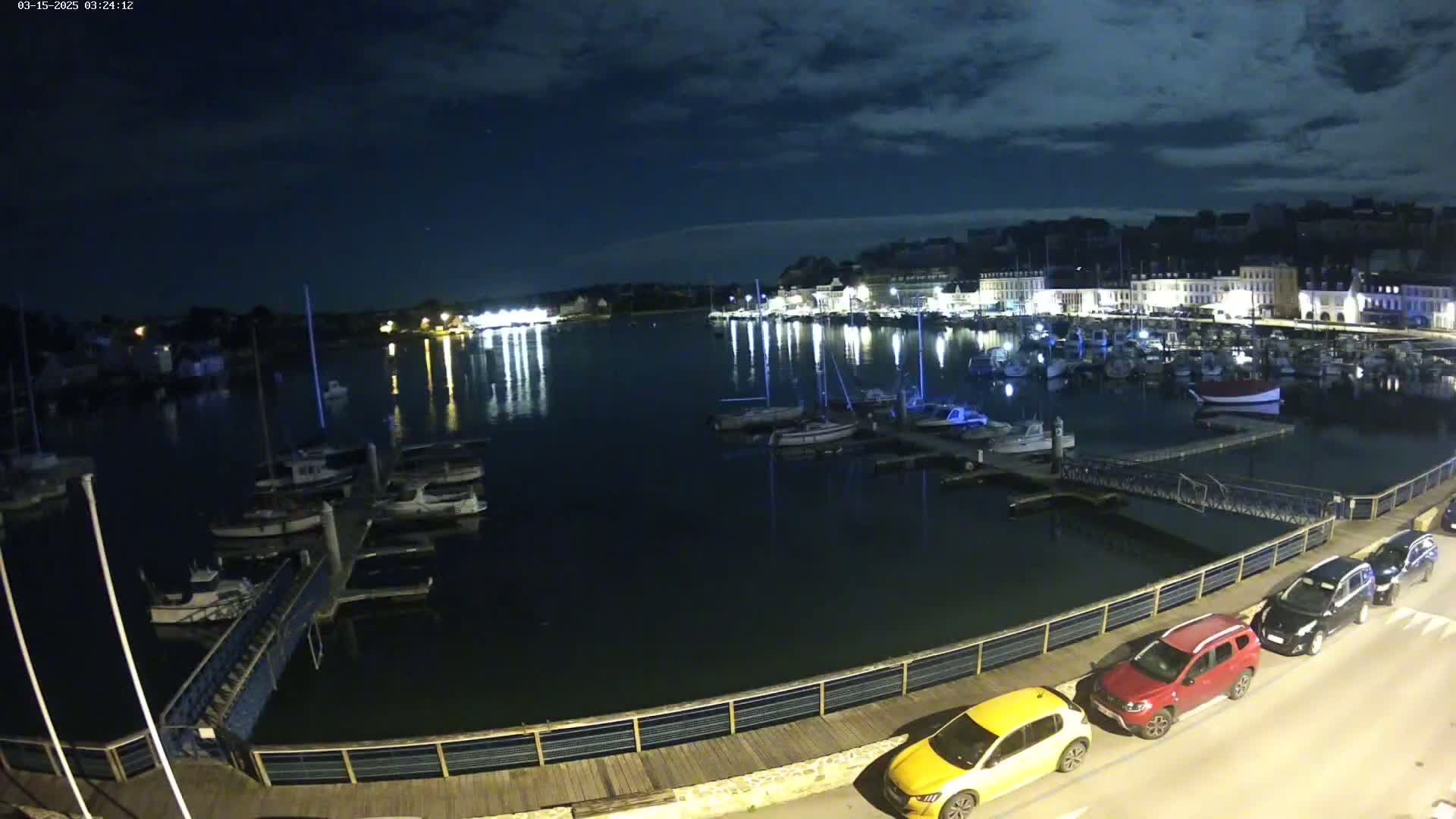 A nighttime view of a harbor filled with boats, docked along a waterfront with cars parked nearby under a partly cloudy sky.
