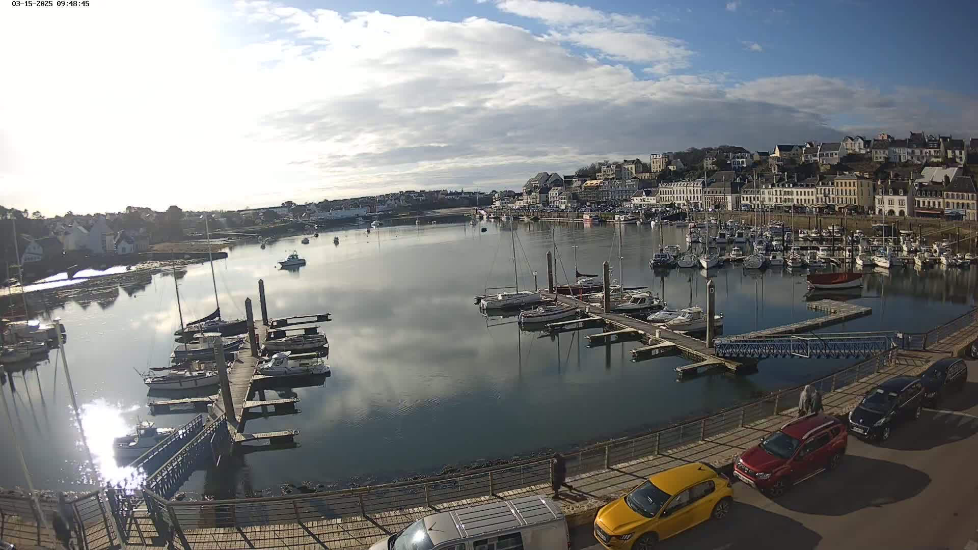 A harbor filled with sailboats and motorboats under a partly cloudy sky is viewed from a waterfront vantage point, with several cars parked along the quay.