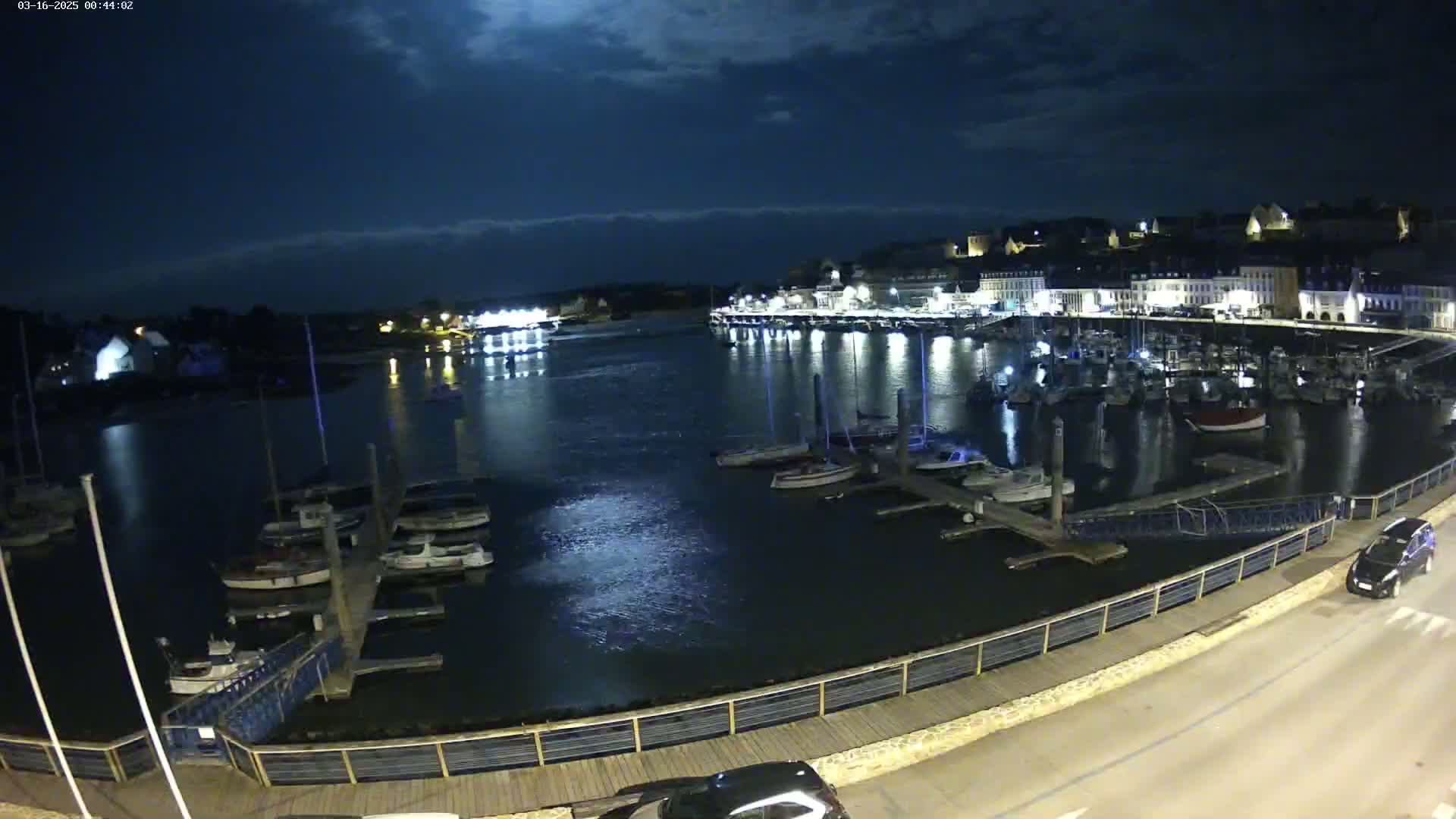 A nighttime harbor scene shows many boats docked along a waterfront, with a town lit up in the background under a mostly cloudy sky.