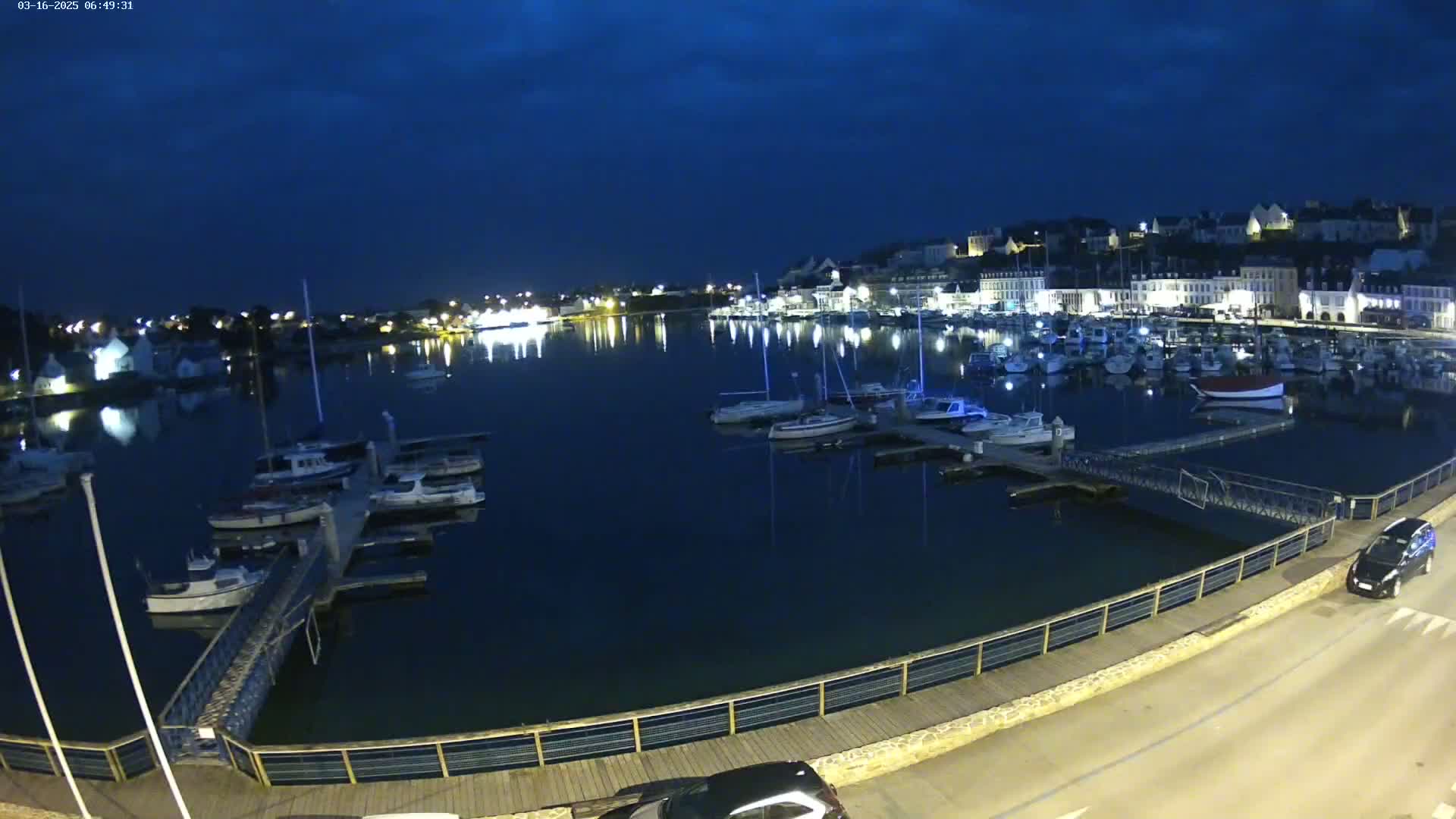 A nighttime view of a harbor filled with boats, with a town illuminated on the opposite shore under a dark, clear sky.