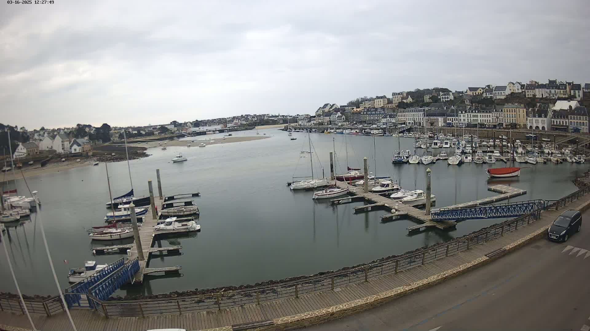 A harbor filled with numerous sailboats and motorboats on a cloudy day, is situated next to a town with buildings lining the waterfront.