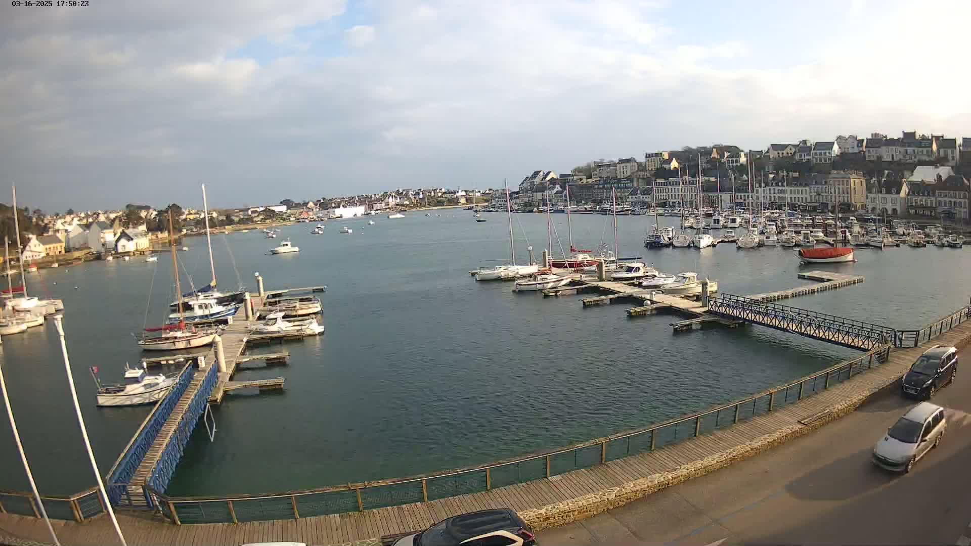 A harbor filled with sailboats and motorboats is nestled beside a town under a partly cloudy sky.