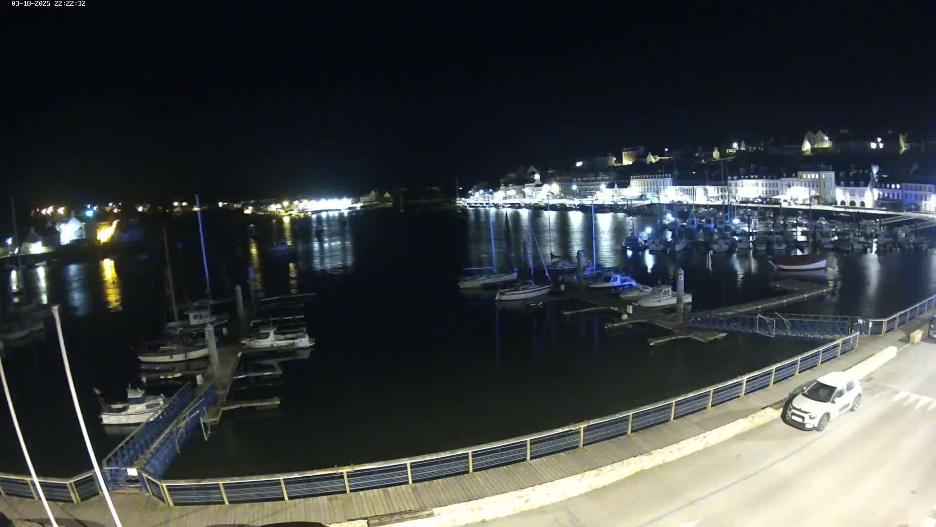 A nighttime view of a harbor filled with boats, docked along a waterfront with buildings illuminated in the background under a clear, dark sky.