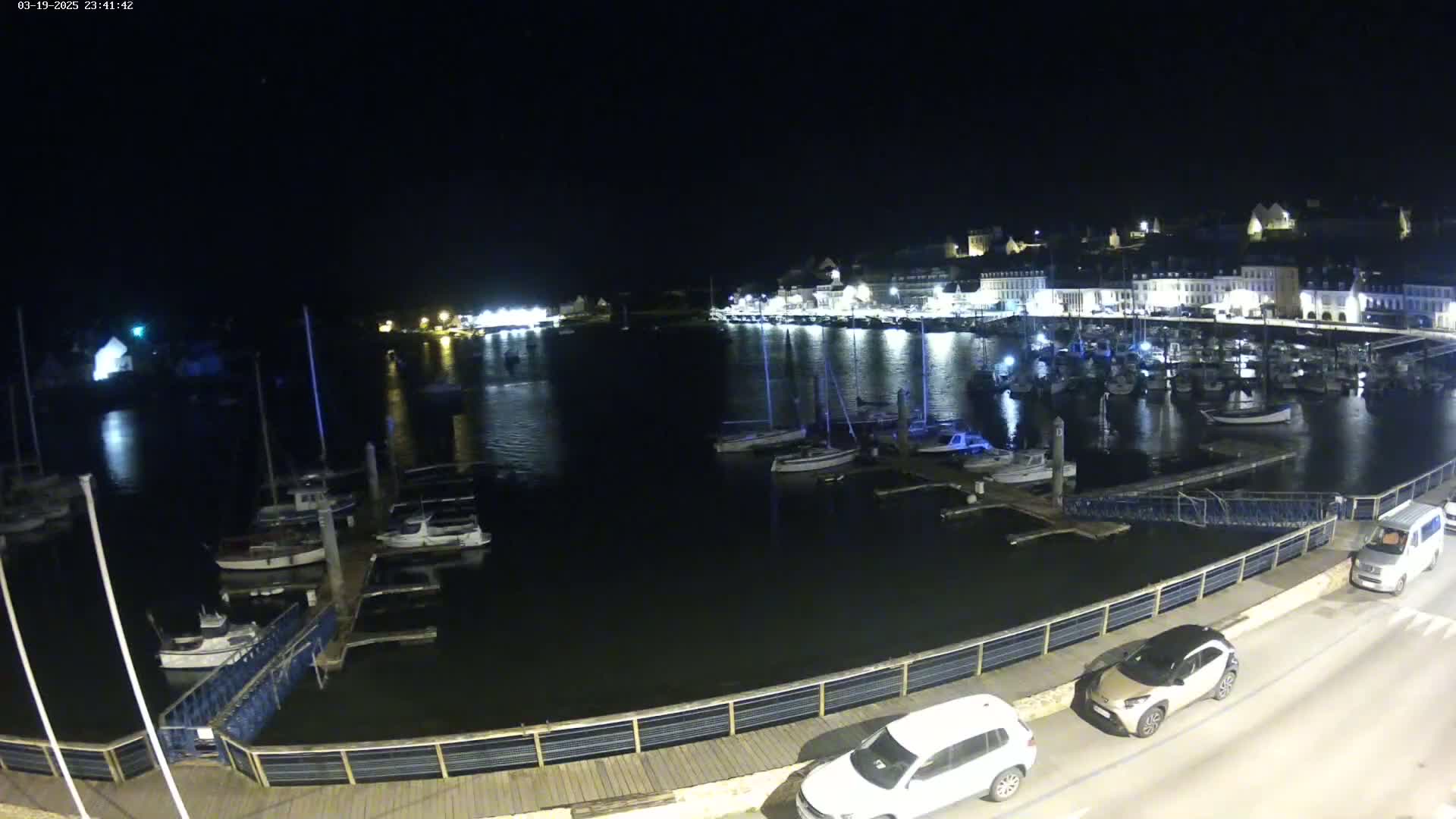 At night, a harbor filled with boats is illuminated, with a town's lights reflecting on the water, and cars parked along a nearby walkway.
