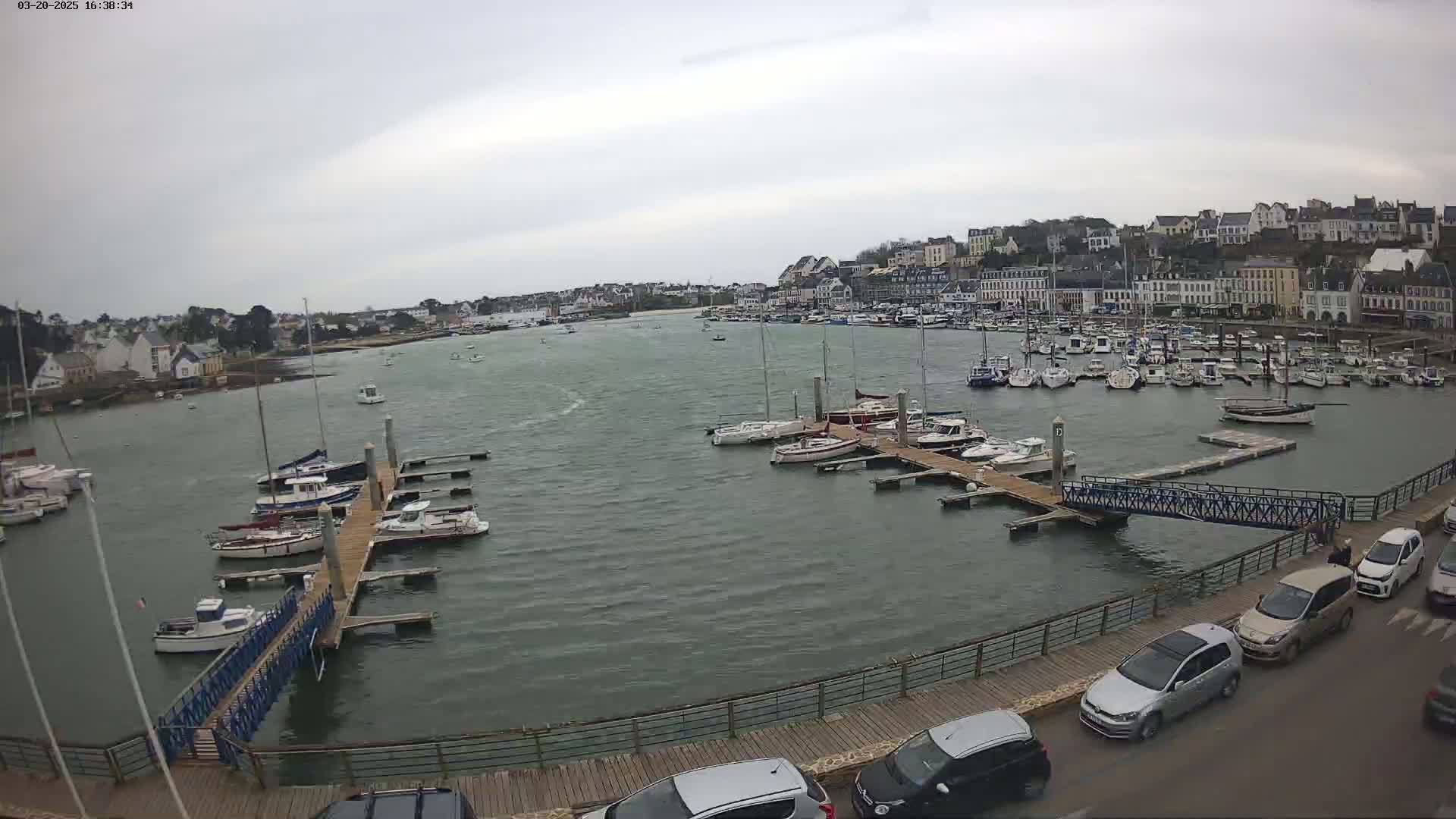 A harbor filled with numerous boats and yachts under an overcast sky, with a town visible in the background and cars parked along a nearby waterfront walkway.